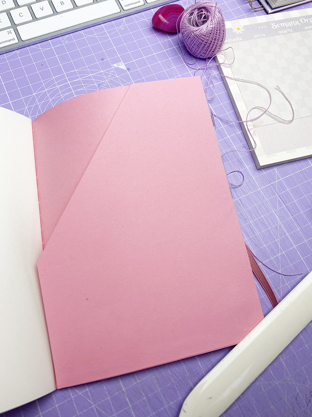 Caderno cor-de-rosa aberto sobre tapete de corte roxo com novelo de linha lilás e régua branca.