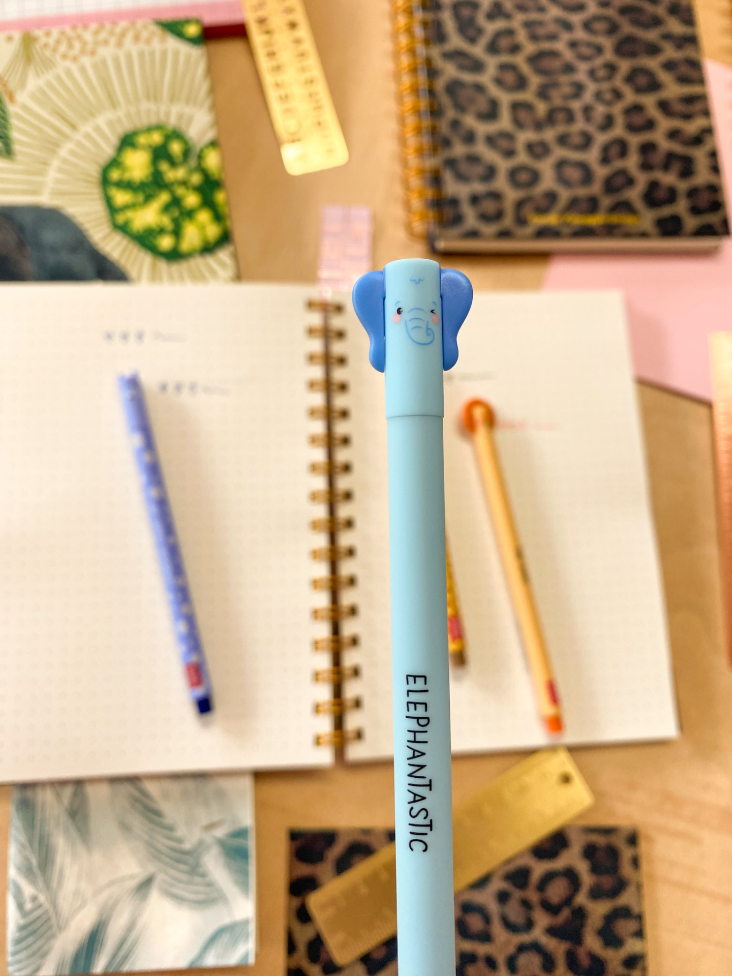 Caneta azul claro com tampa de elefante e texto ELEPHANTASTIC sobre mesa com cadernos e réguas