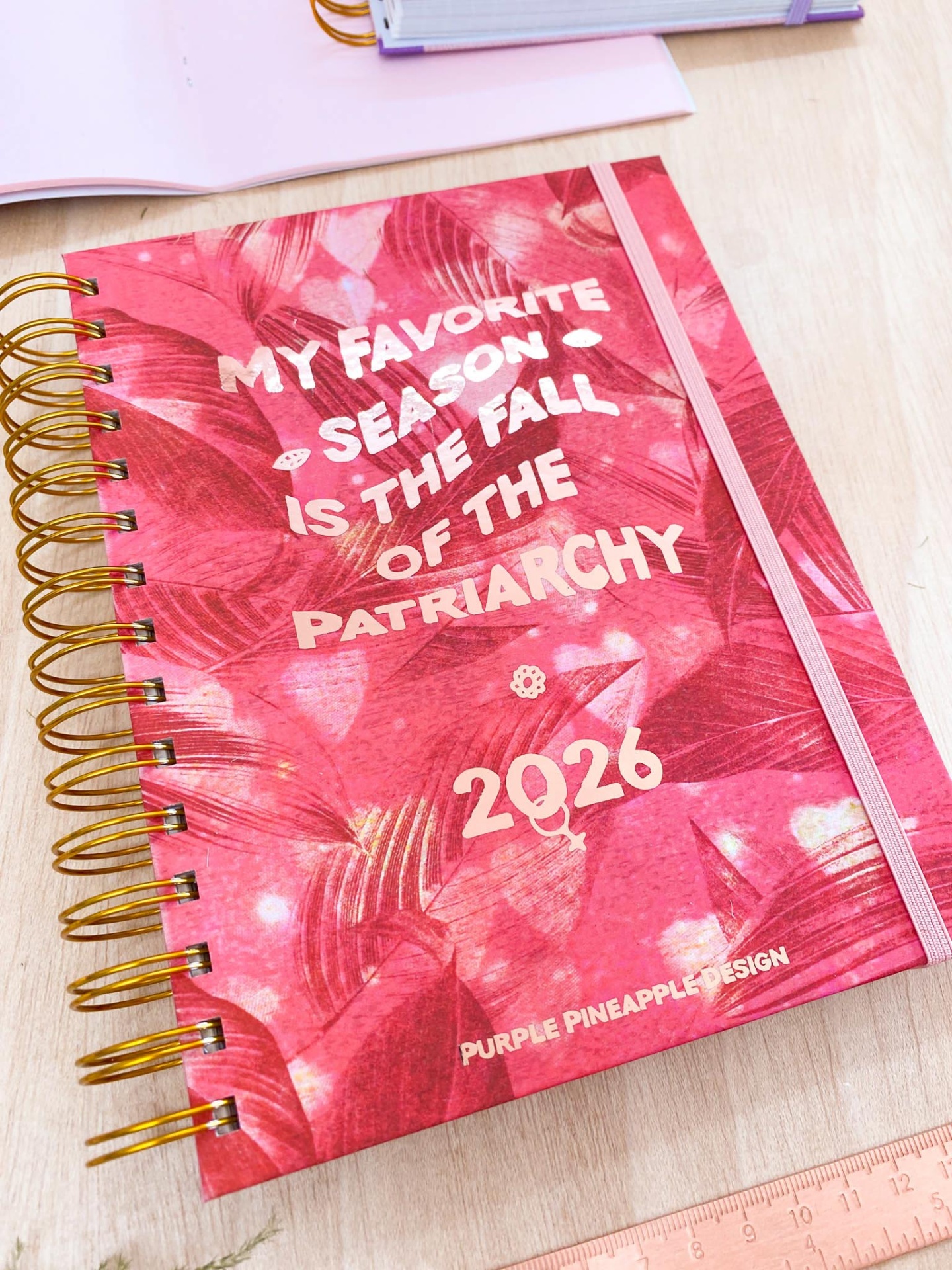 Caderno rosado com texto branco e espiral dourada numa mesa de madeira