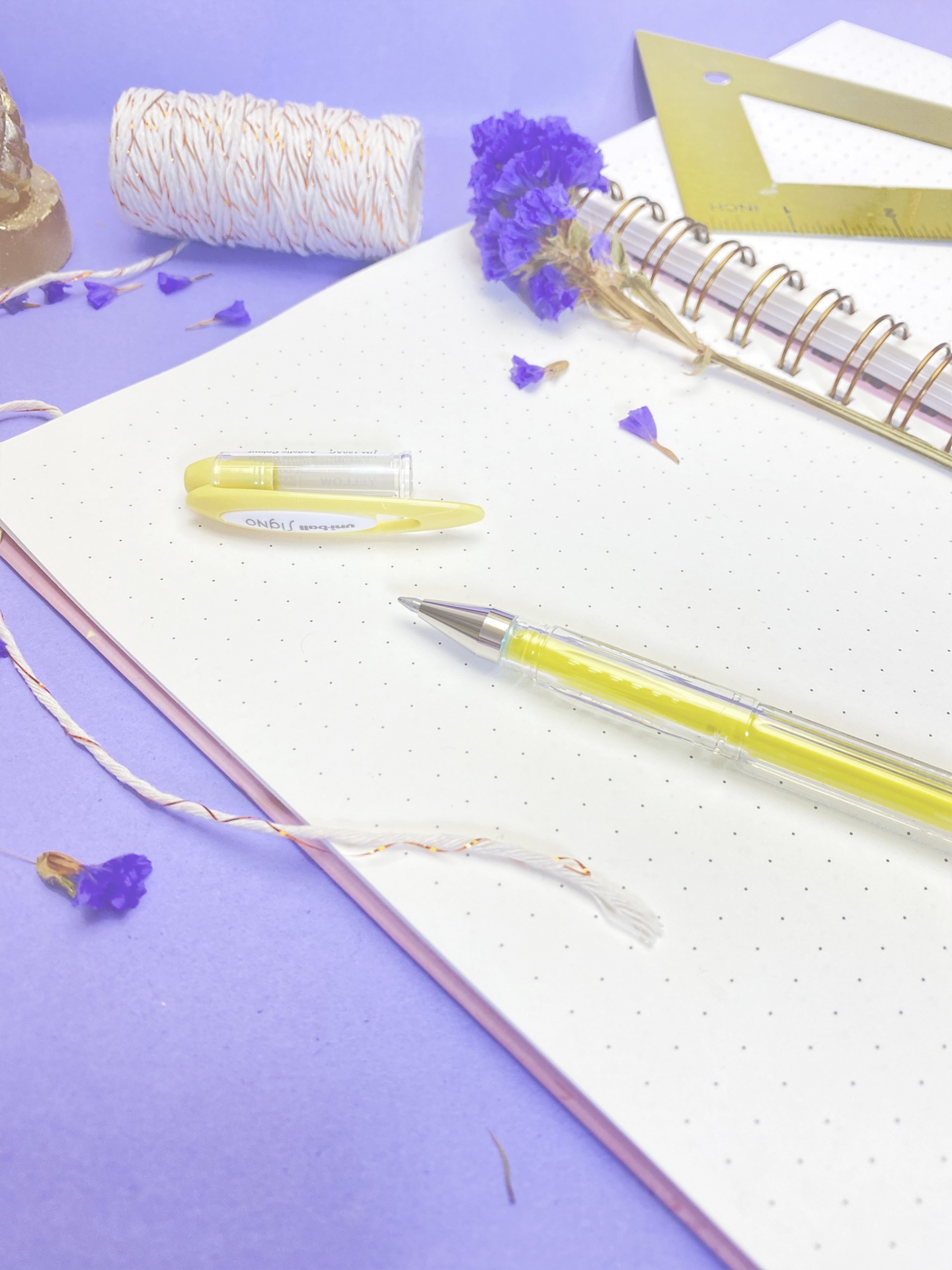 Caderno aberto com caneta amarela, flores secas e acessórios de papelaria sobre fundo lilás