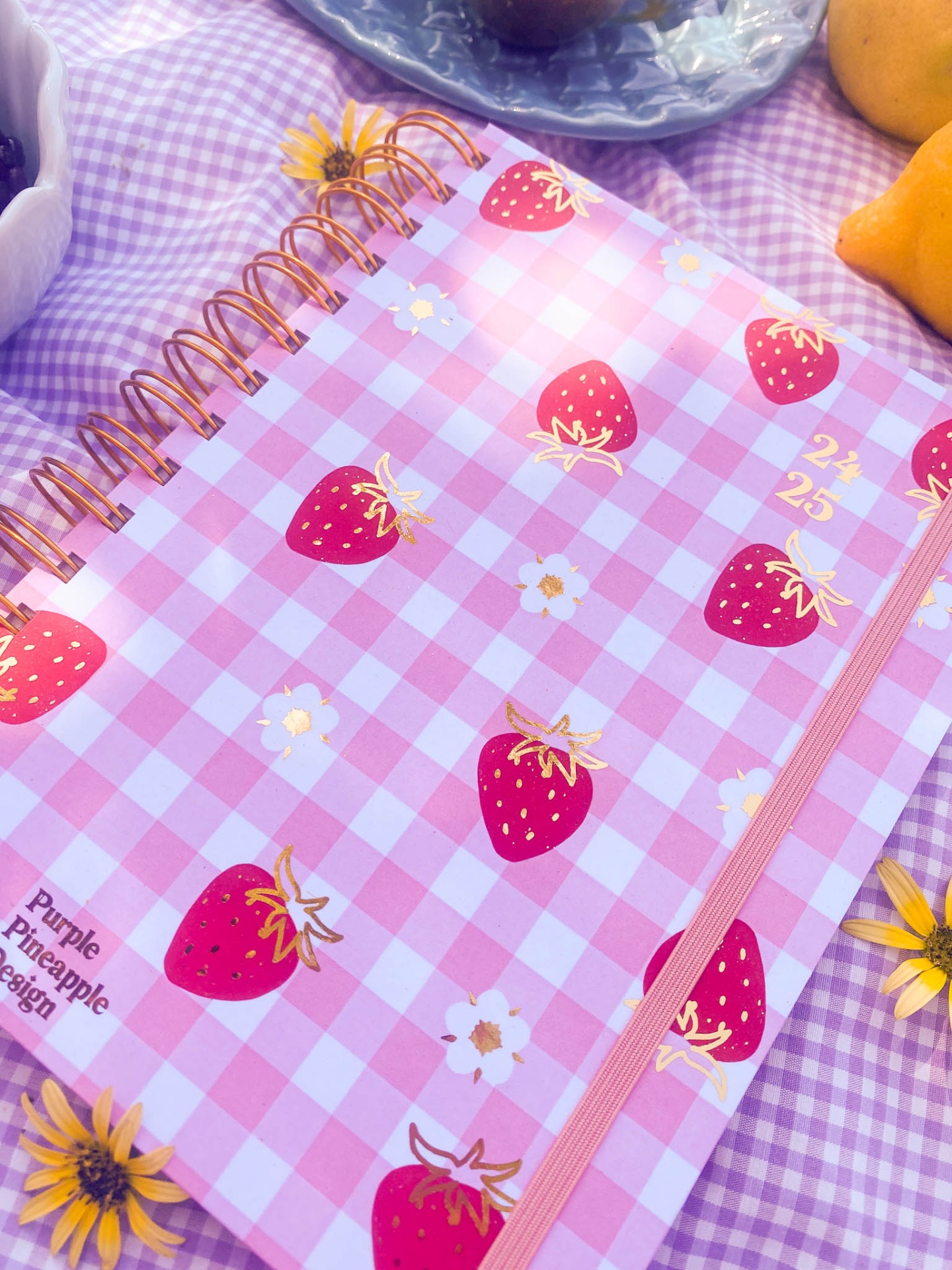 Caderno com capa xadrez rosa e branco com morangos e flores, espiral dourada e banda elástica rosa