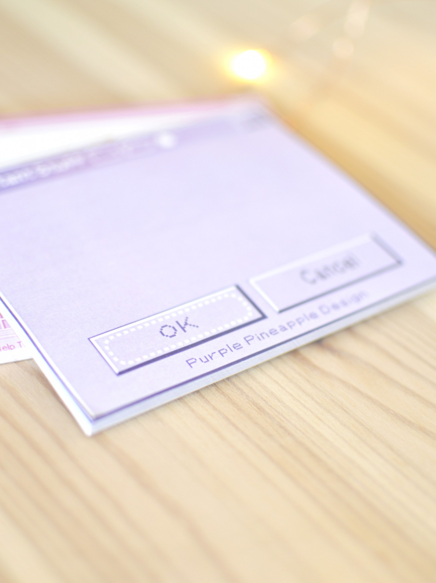 Cartão lilás com texto 'OK', 'Cancel' e 'Purple Pineapple Design' em mesa de madeira