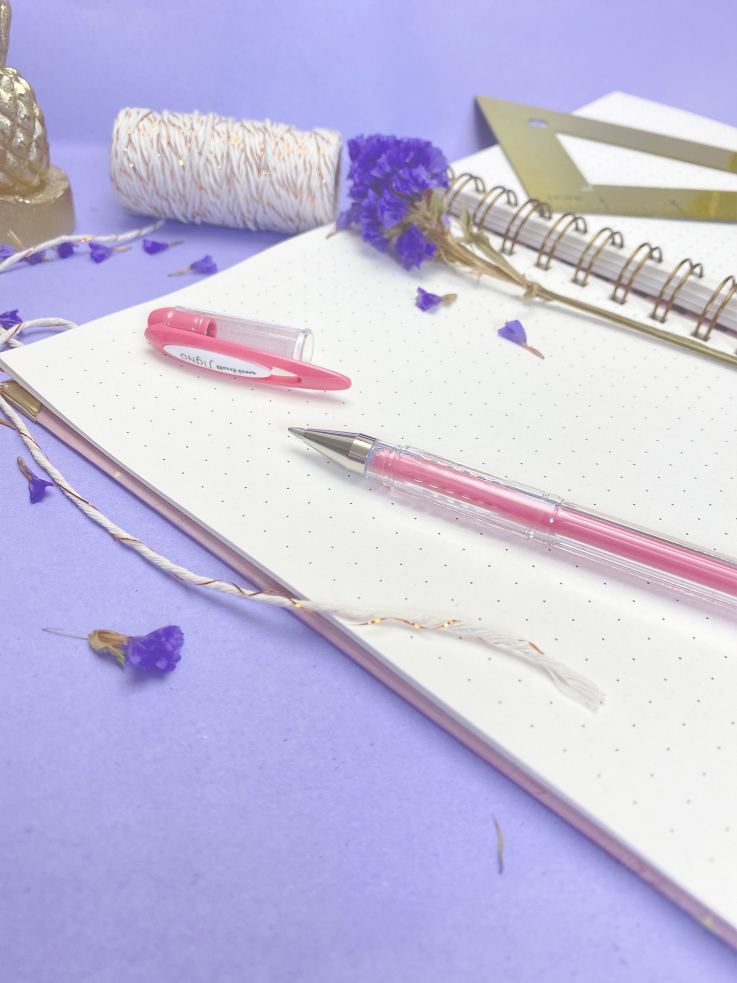 Caderno aberto com caneta rosa, flores secas e material de escritório sobre superfície lilás