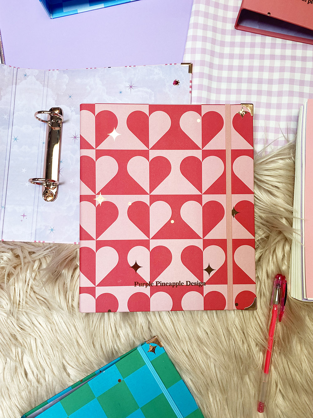 Caderno rosa e vermelho com corações e argolas metálicas sobre tapete felpudo com caneta rosa ao lado