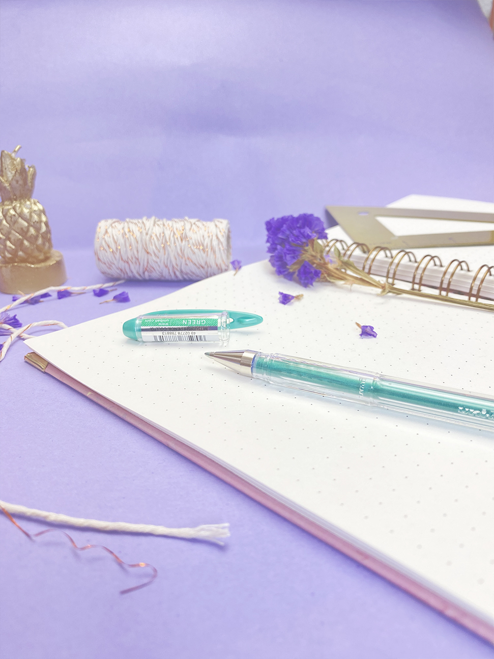 Caderno aberto com caneta verde transparente, flor seca roxa e rolo de fio branco com dourado sobre fundo lilás
