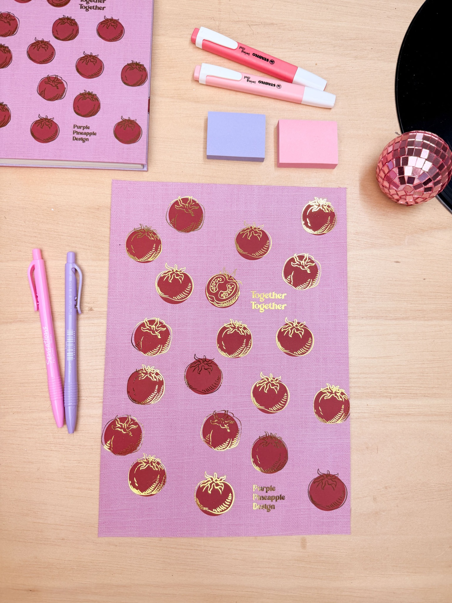 Folha decorativa roxa com desenhos de tomates vermelhos, texto dourado e objetos de papelaria coloridos sobre mesa de madeira.
