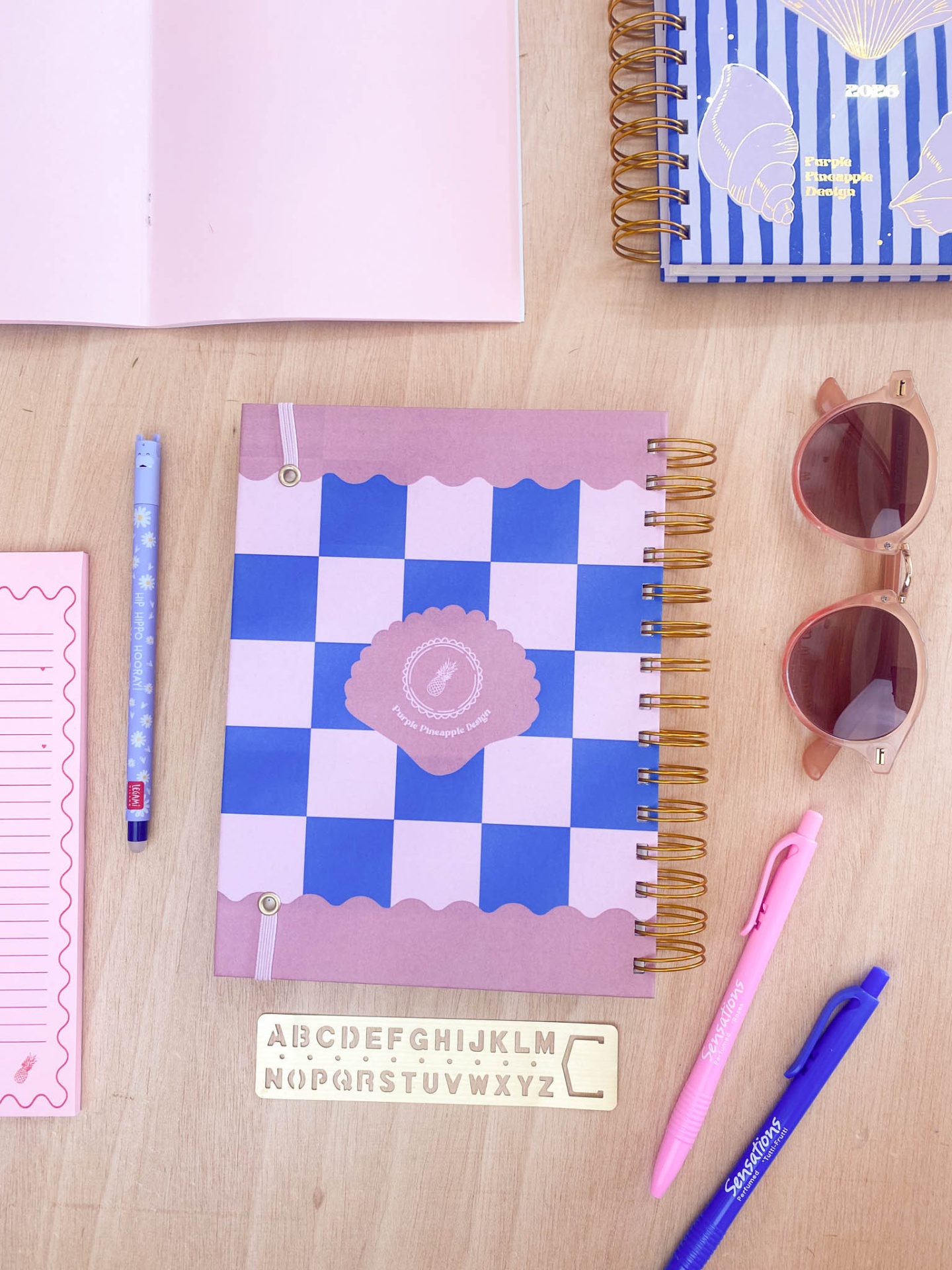 Caderno espiral lilás e azul xadrez com canetas, régua alfabética, óculos de sol rosa e bloco de notas rosa sobre mesa de madeira