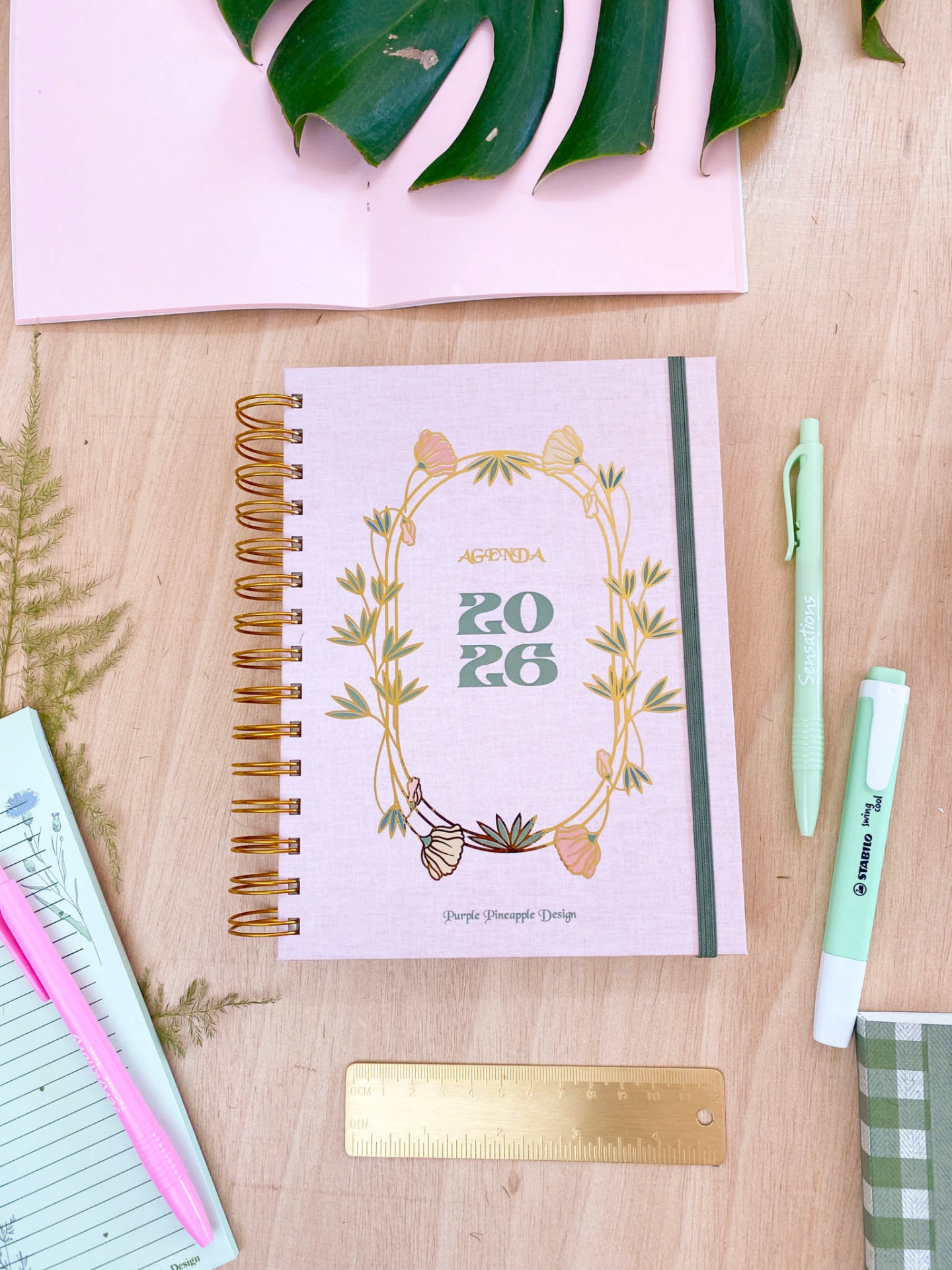 Agenda 2026 com capa rosa e ilustração floral, espiral dourada e elástico cinza numa mesa de madeira com canetas e régua