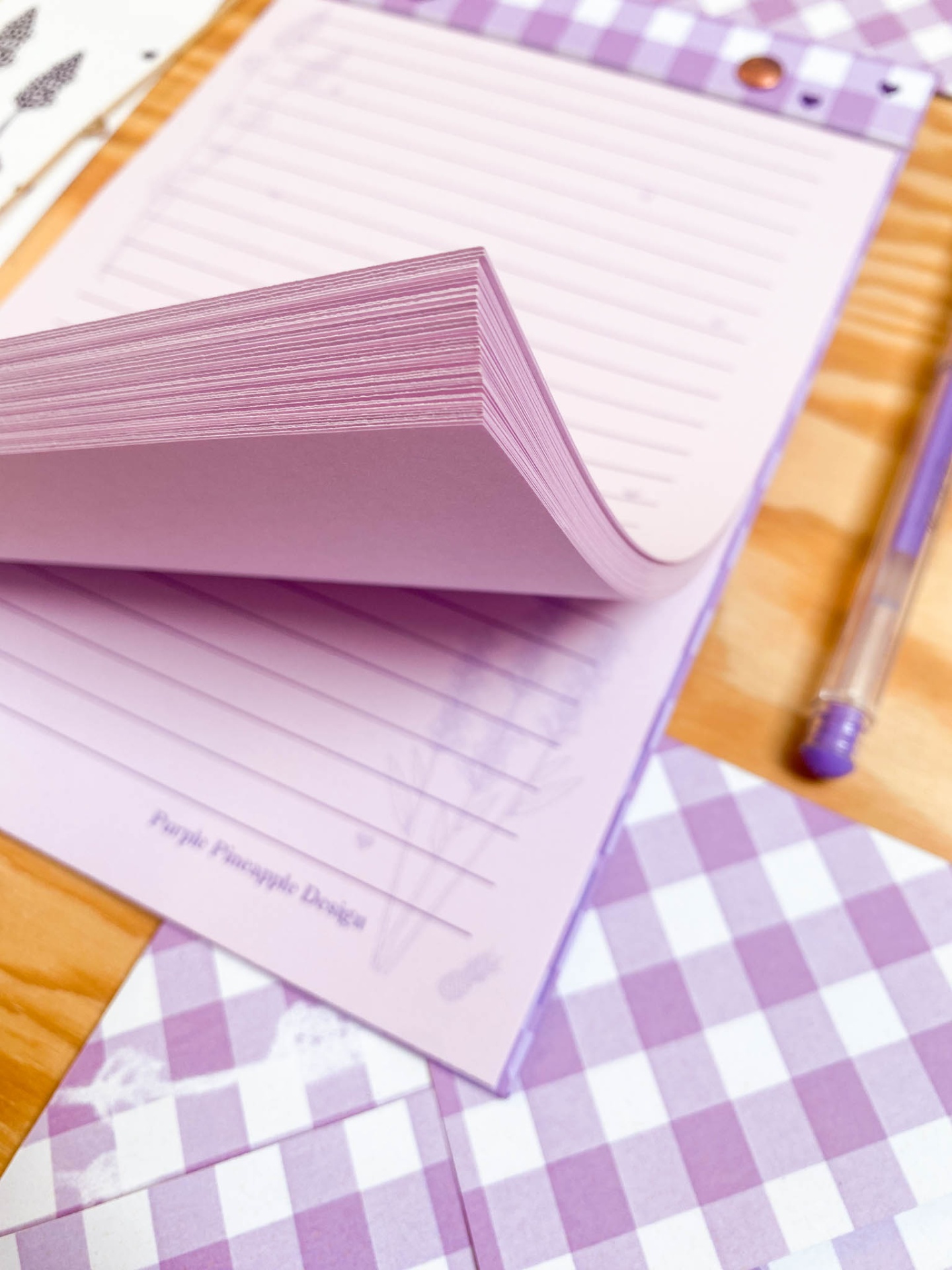 bloco de notas rosa, caneta lilás e envelopes xadrez lilás e branco sobre mesa de madeira