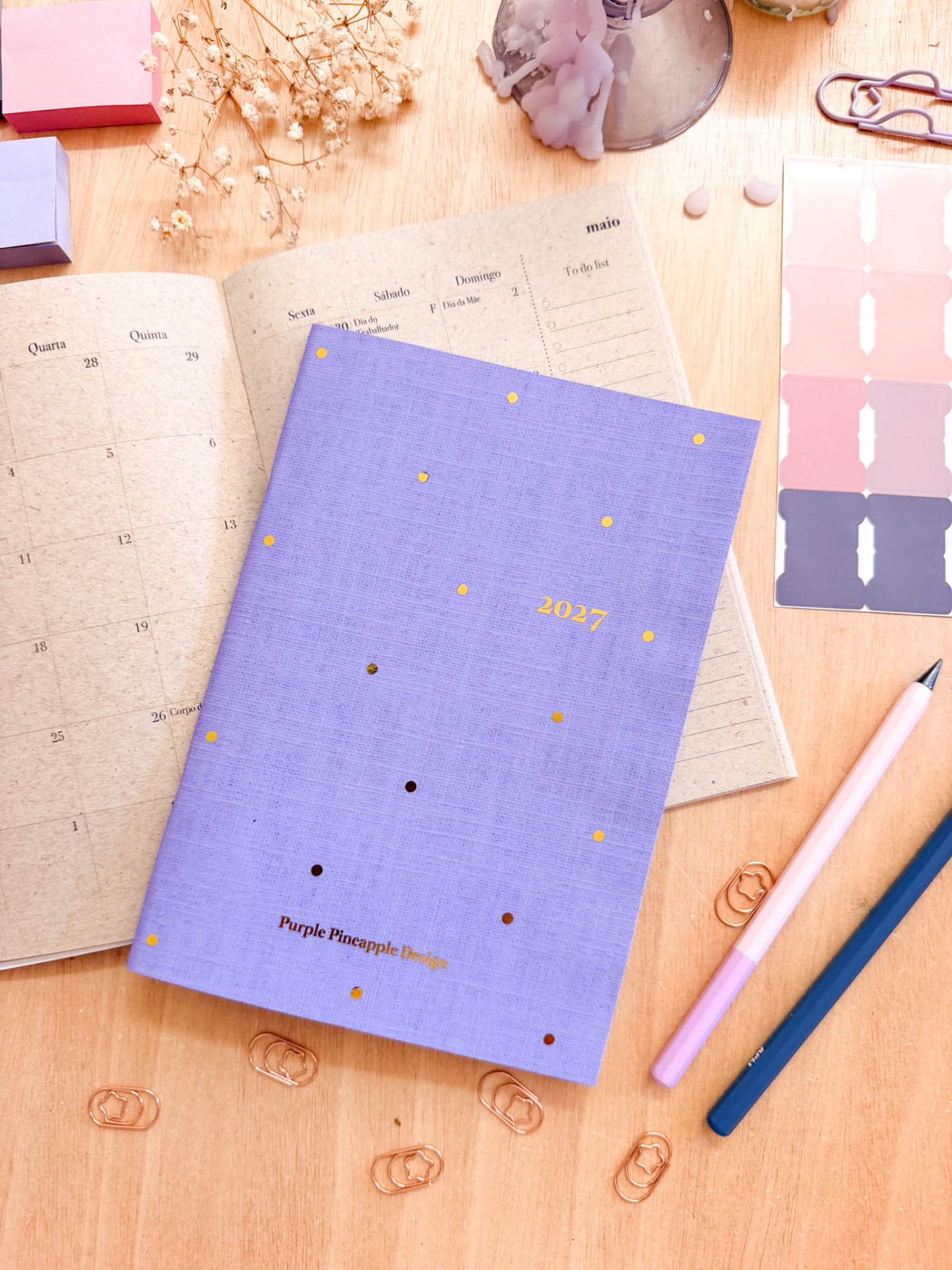 Agenda anual com capa roxa e pontos dourados sobre mesa com calendário, canetas e clips