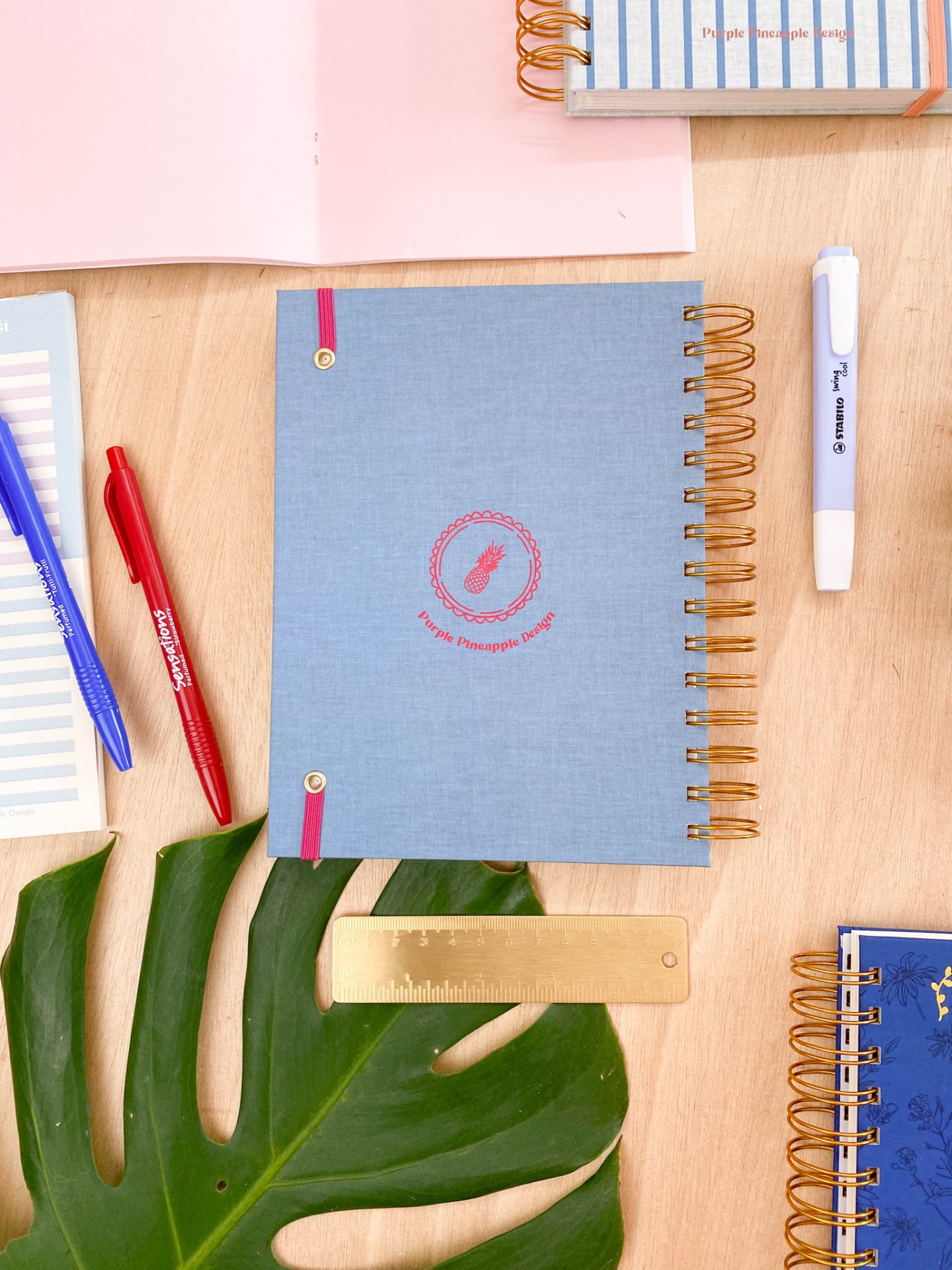Caderno azul com capa de tecido e texto Purple Pineapple Design sobre mesa de madeira com canetas, régua e planta verde.
