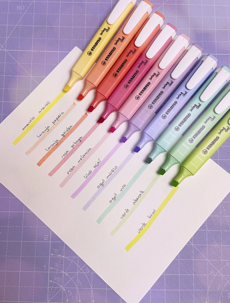 Sete marcadores Stabilo de cores pastel com linhas e anotações em folha branca sobre superfície quadriculada lilás