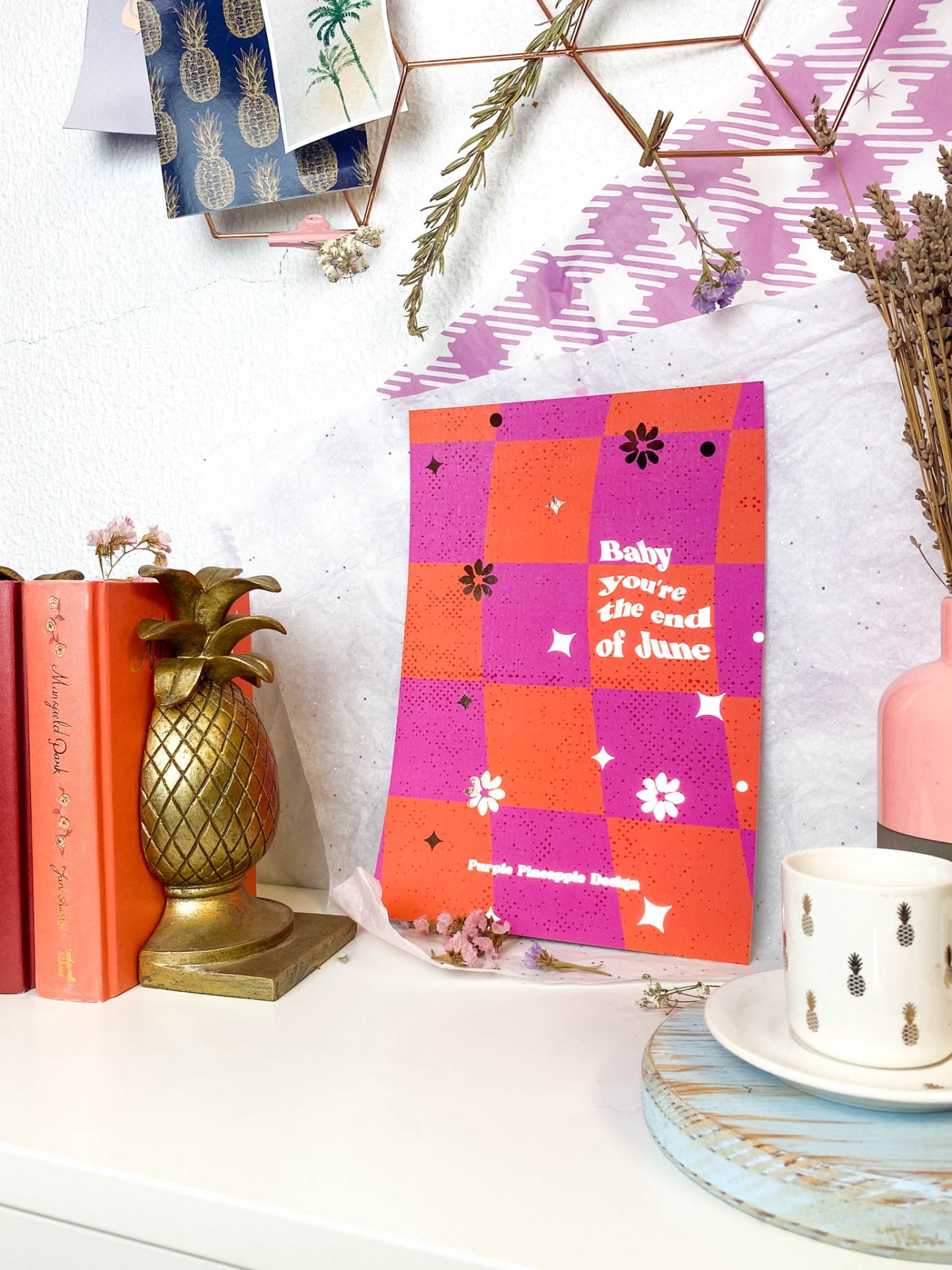 Cartaz colorido com texto e padrão floral numa mesa com livros, suporte abacaxi dourado e chávena com pires azul.