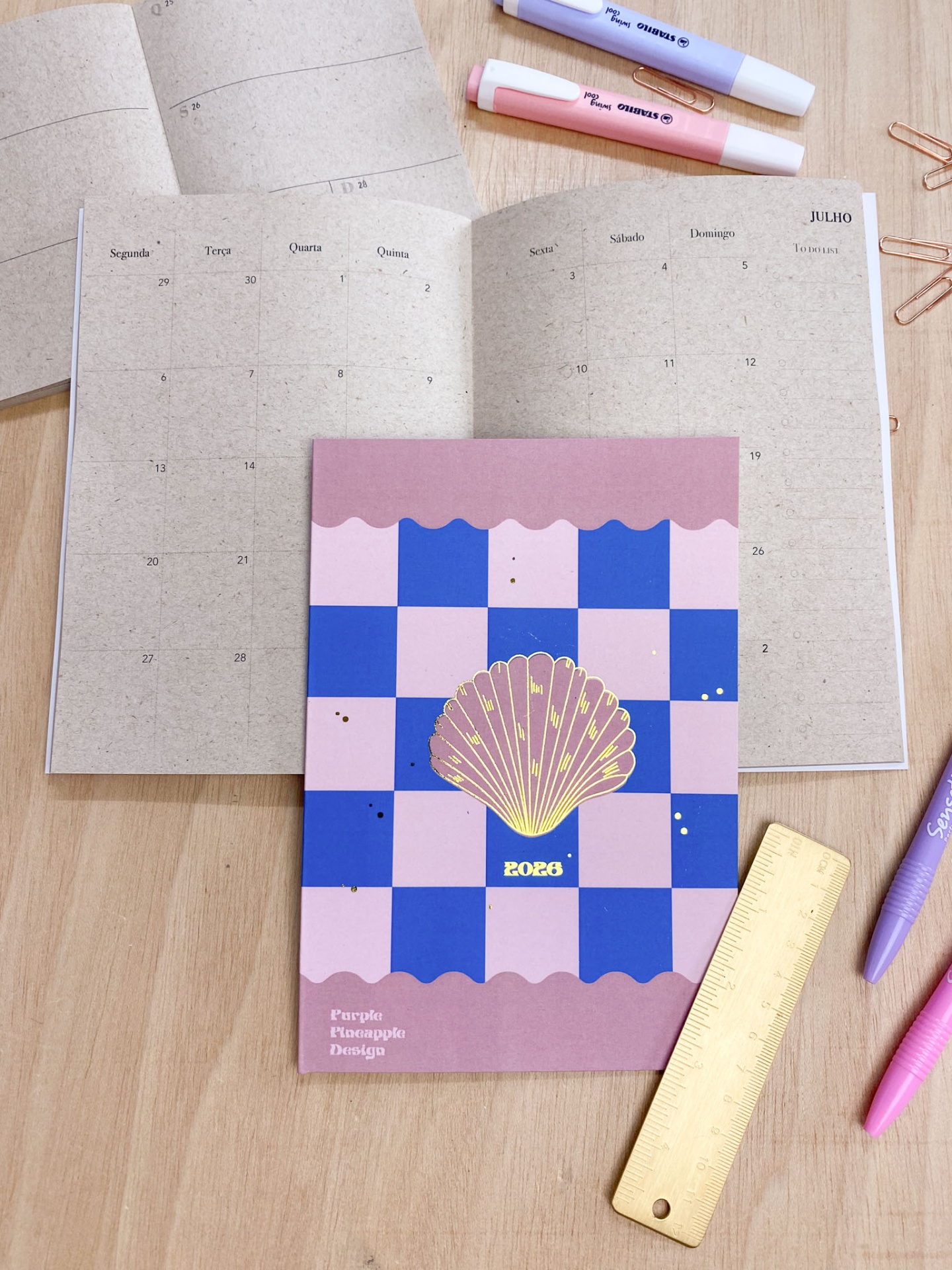 Agenda com capa xadrez azul e rosa com concha dourada e texto 2023, aberta em julho, sobre mesa com canetas, clips e régua.