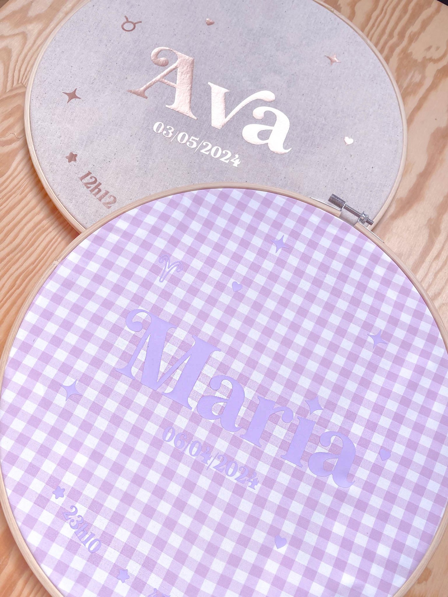 Dois quadros redondos bordados com nomes Ava e Maria e datas em tecido lilás xadrez e cinza claro sobre madeira