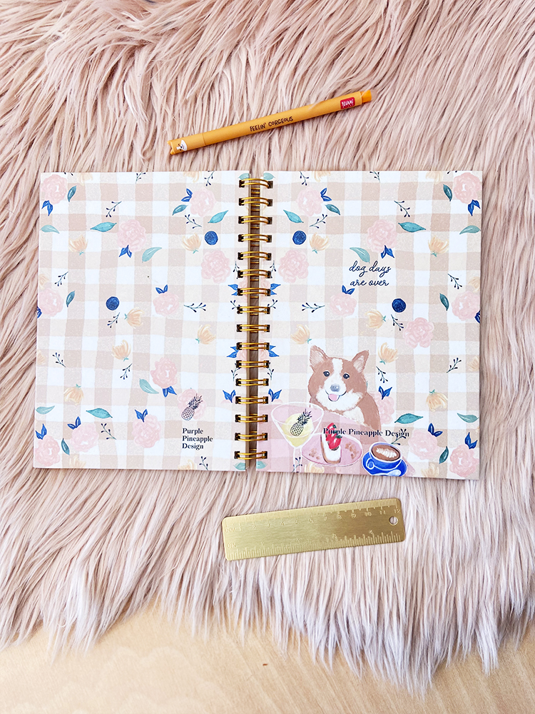 Caderno espiral aberto com ilustração de cão e padrão floral, régua dourada e caneta amarela sobre superfície rosa felpuda