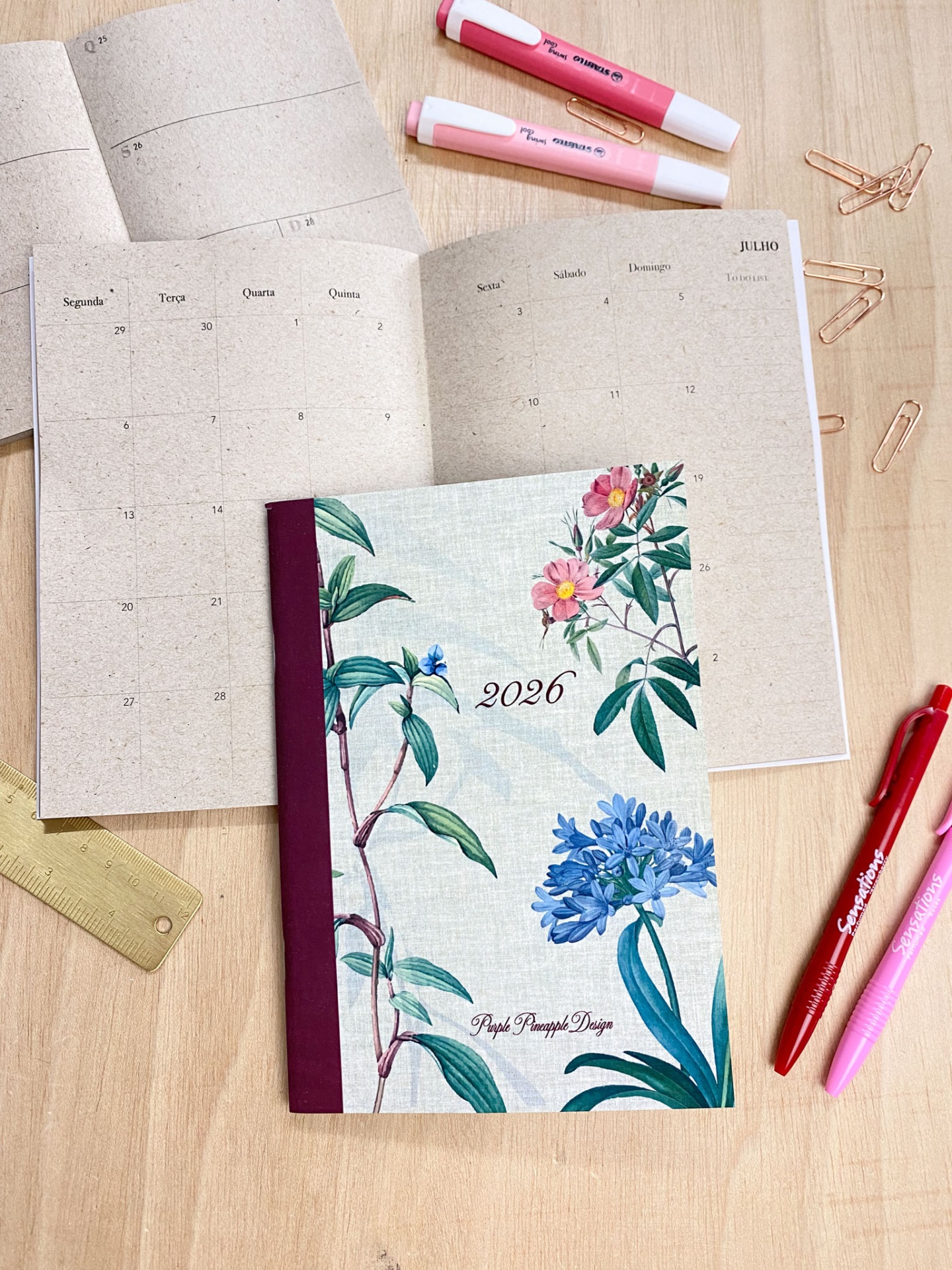 Agenda 2026 com capa floral e calendário aberto sobre mesa com material de escrita.