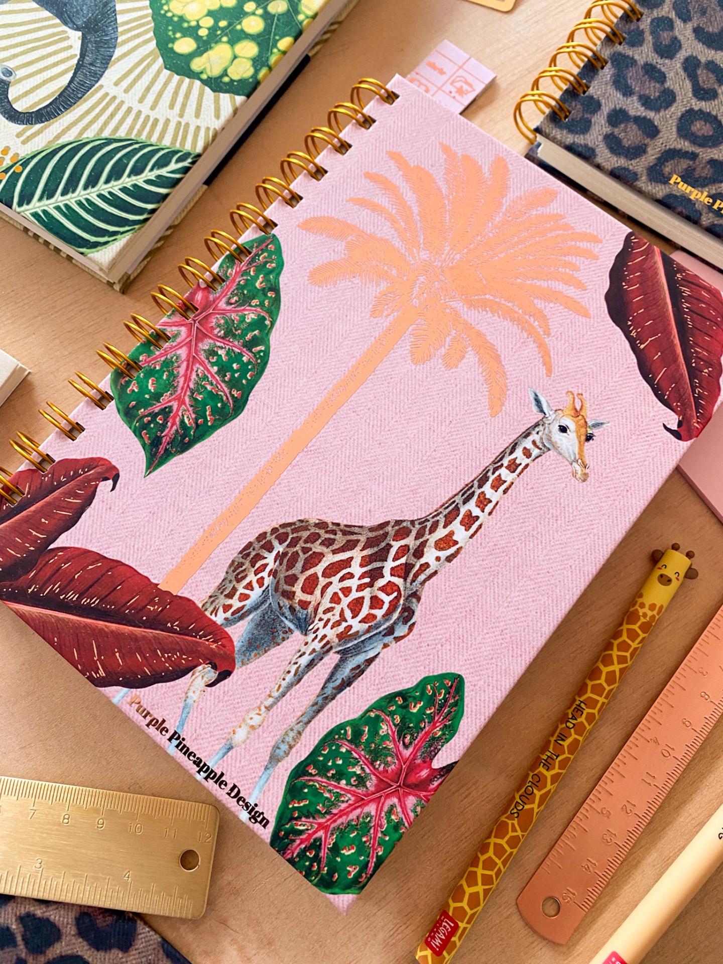 Caderno rosa com girafa e folhas, régua dourada e lápis de girafa
