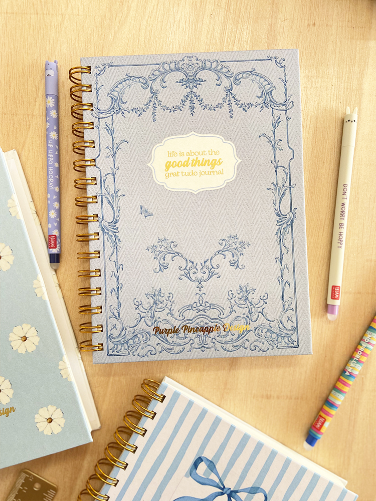 Agenda ou caderno com capa cinzenta decorada e texto em dourado, rodeado por marcadores e outros cadernos sobre mesa de madeira.