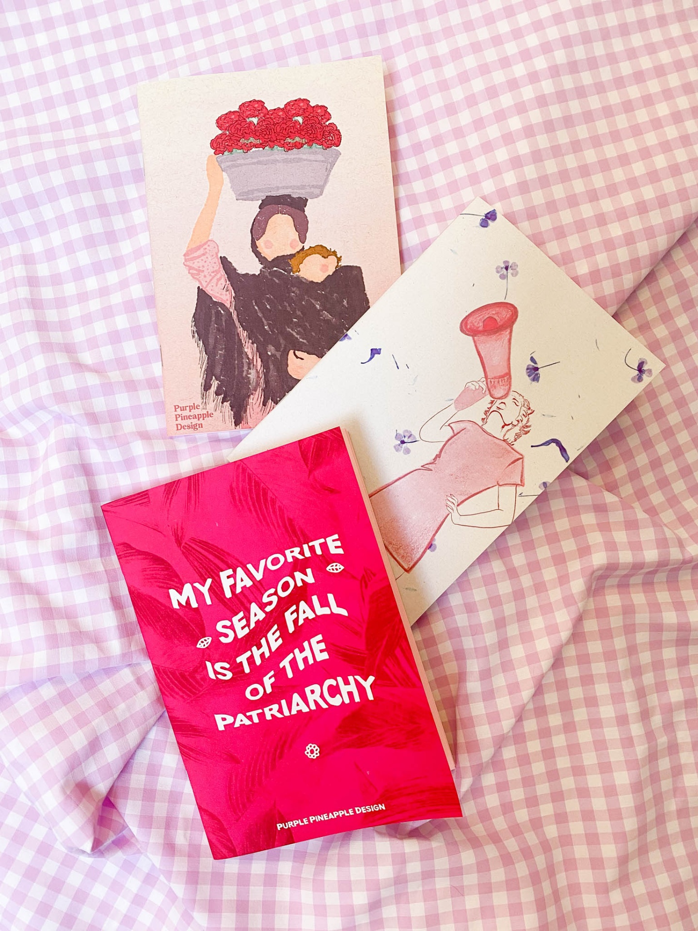 Três postais coloridos sobre tecido xadrez lilás e branco com texto político e ilustrações femininas.