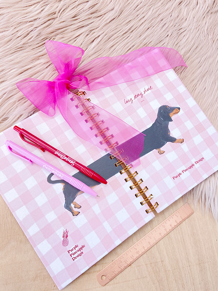 Caderno rosa às quadrados com desenho de dachshund, canetas e régua