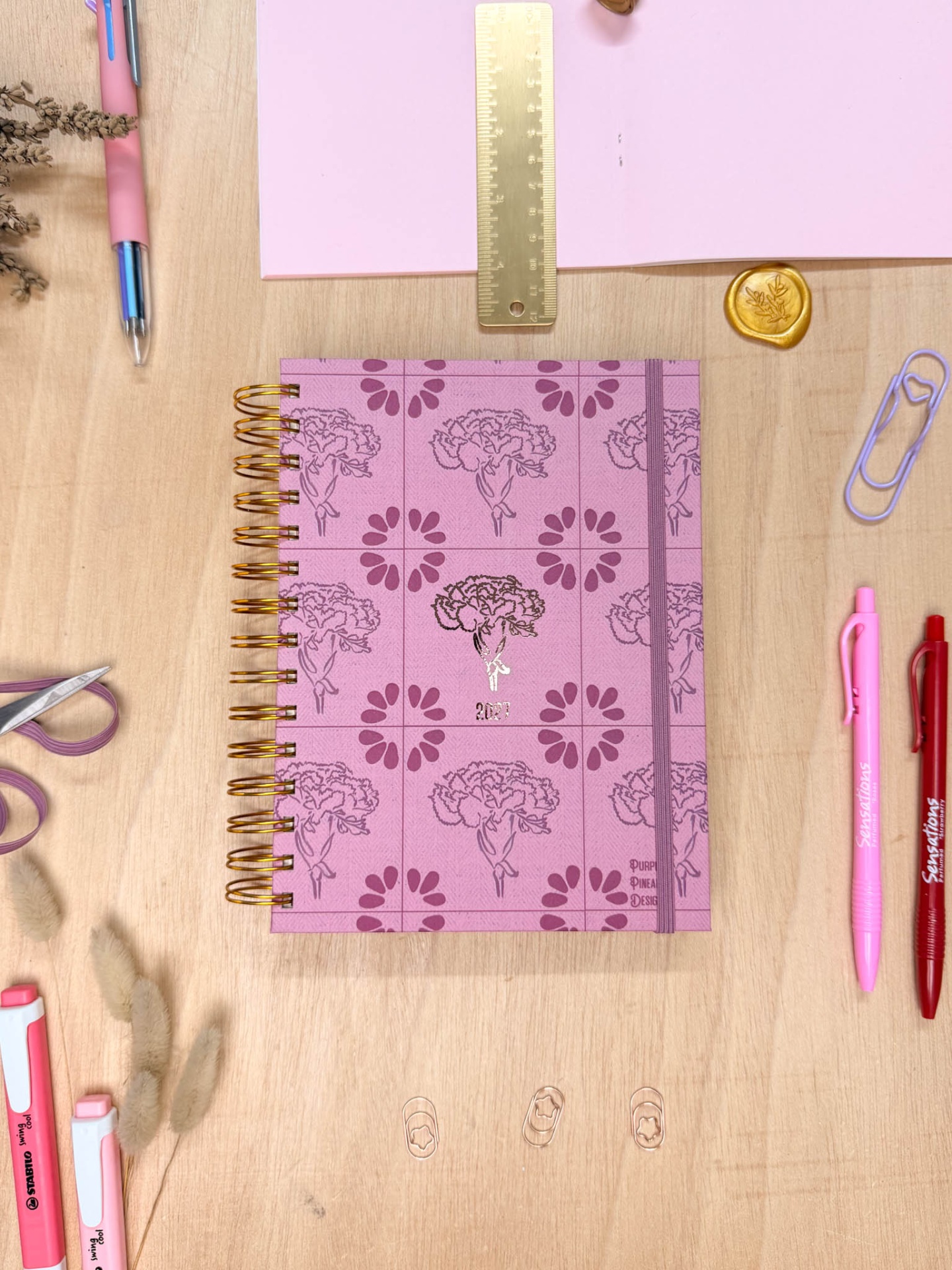 Caderno rosa floral espiral dourada com régua, canetas e clips numa mesa de madeira clara