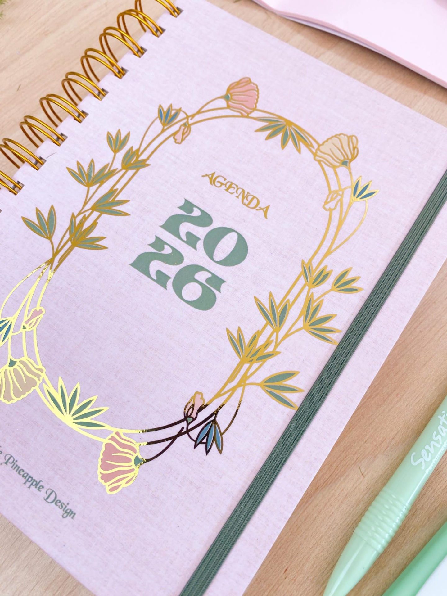 Agenda 2026 rosa clara com detalhes florais dourados e verdes, espiral dourado e fecho elástico verde sobre madeira clara