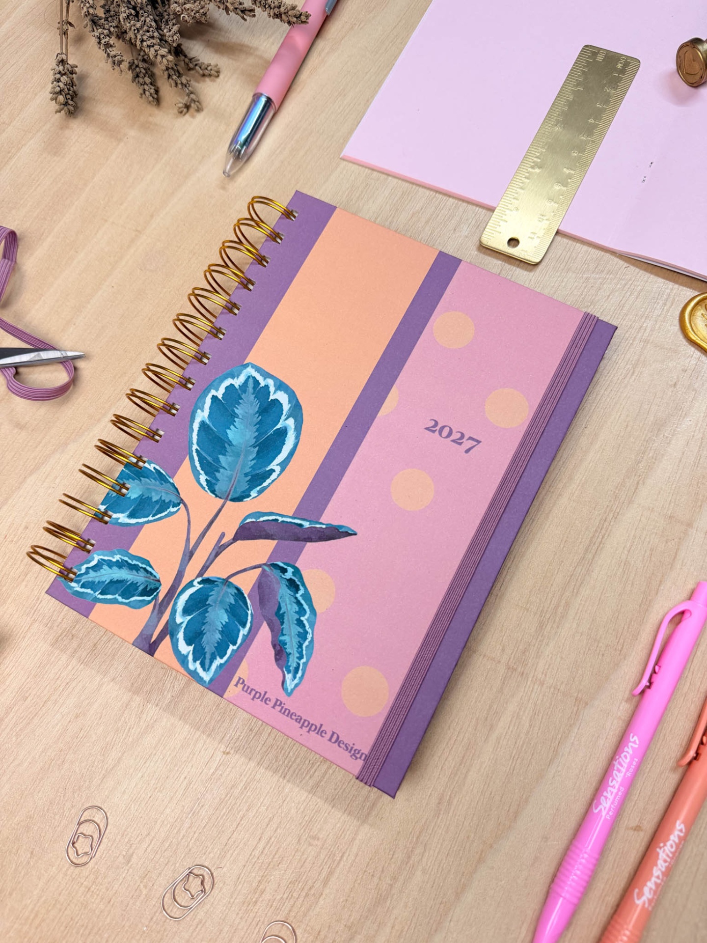 Agenda rosa e lilás com estampa de folhas azuis, canetas rosa e régua dourada sobre mesa de madeira.