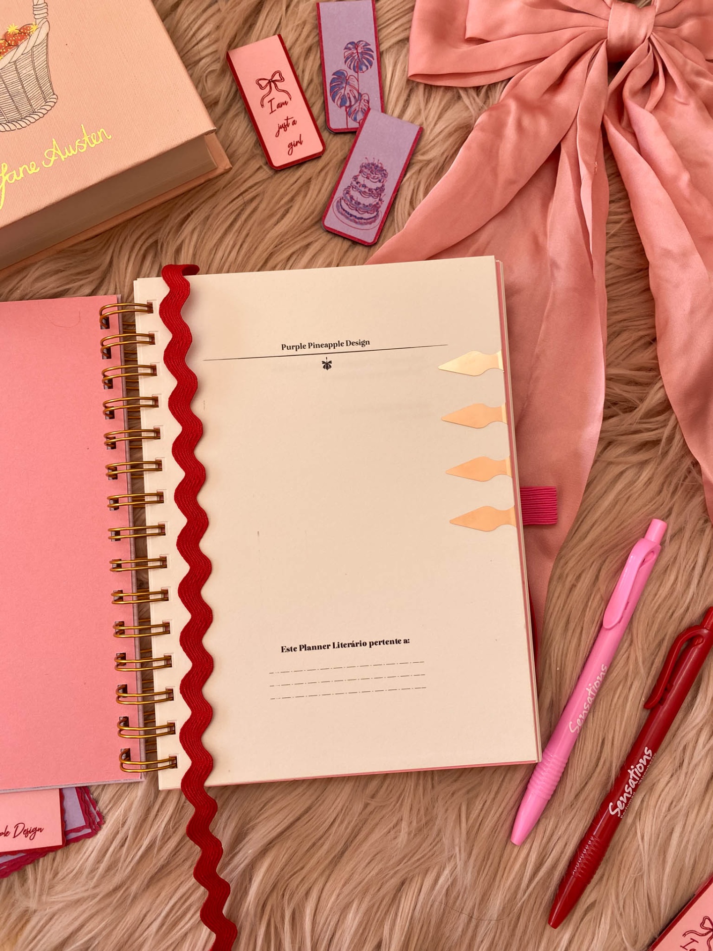 caderno rosa aberto com fitas vermelhas e marcadores, canetas rosa e vermelha, lenço rosa, marcadores de livro rosa e lilás sobre superfície felpuda