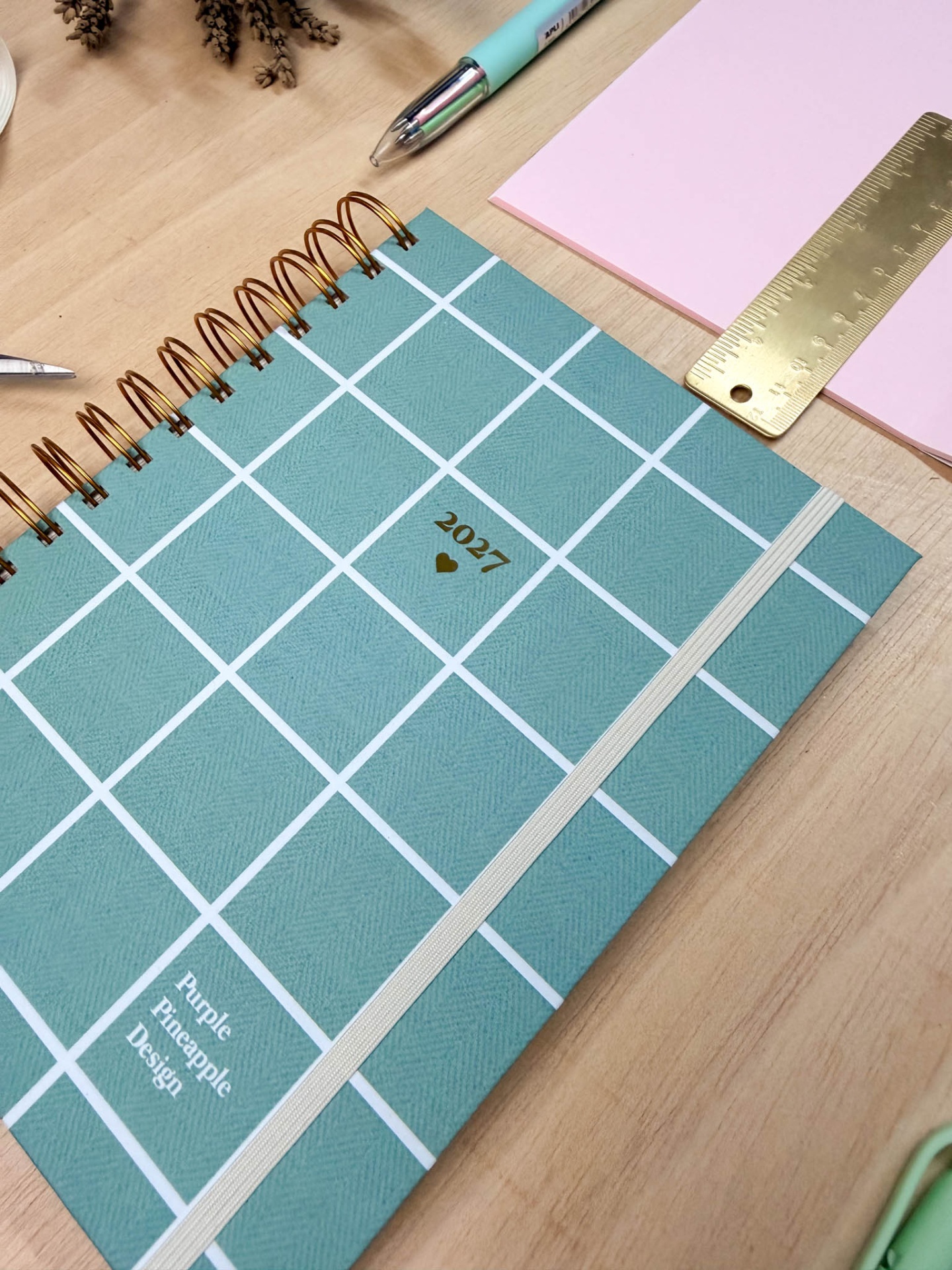 Agenda azul com padrão de quadros brancos e texto 2027 sobre mesa com régua dourada, bloco rosa e caneta azul.