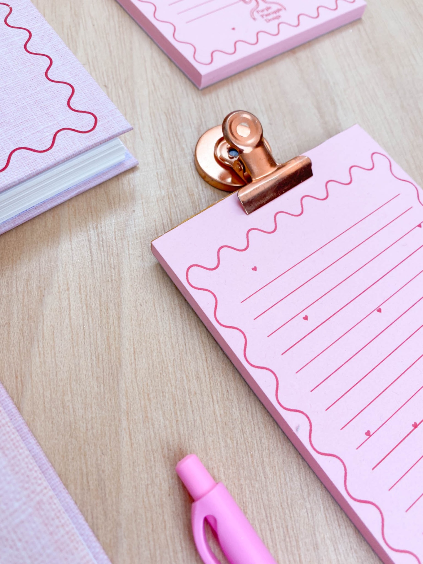 Conjunto de papelaria rosa com bloco de notas, caneta e caderno sobre mesa de madeira