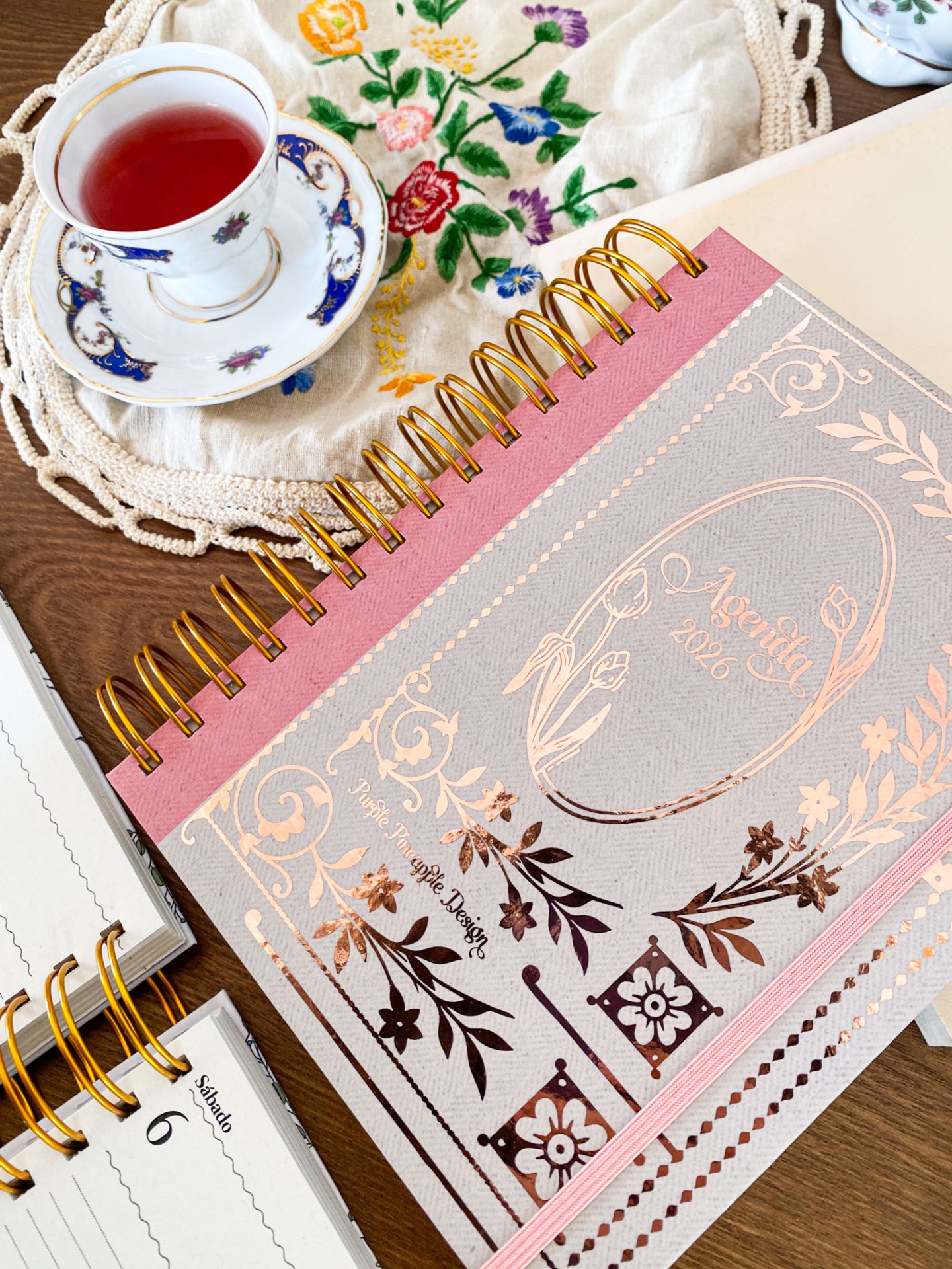 Agenda 2026 com desenho floral e espiral dourada junto a chávena de chá em porcelana e toalha bordada colorida.