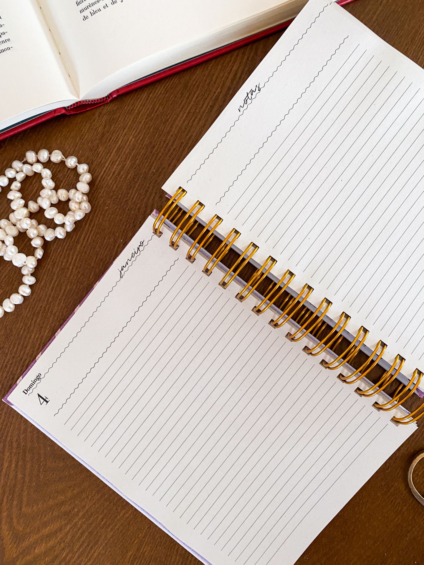 Caderno espiral aberto com texto 'Domingo 4', 'janeiro' e 'notes' numa mesa de madeira junto a livro aberto e colar de pérolas.