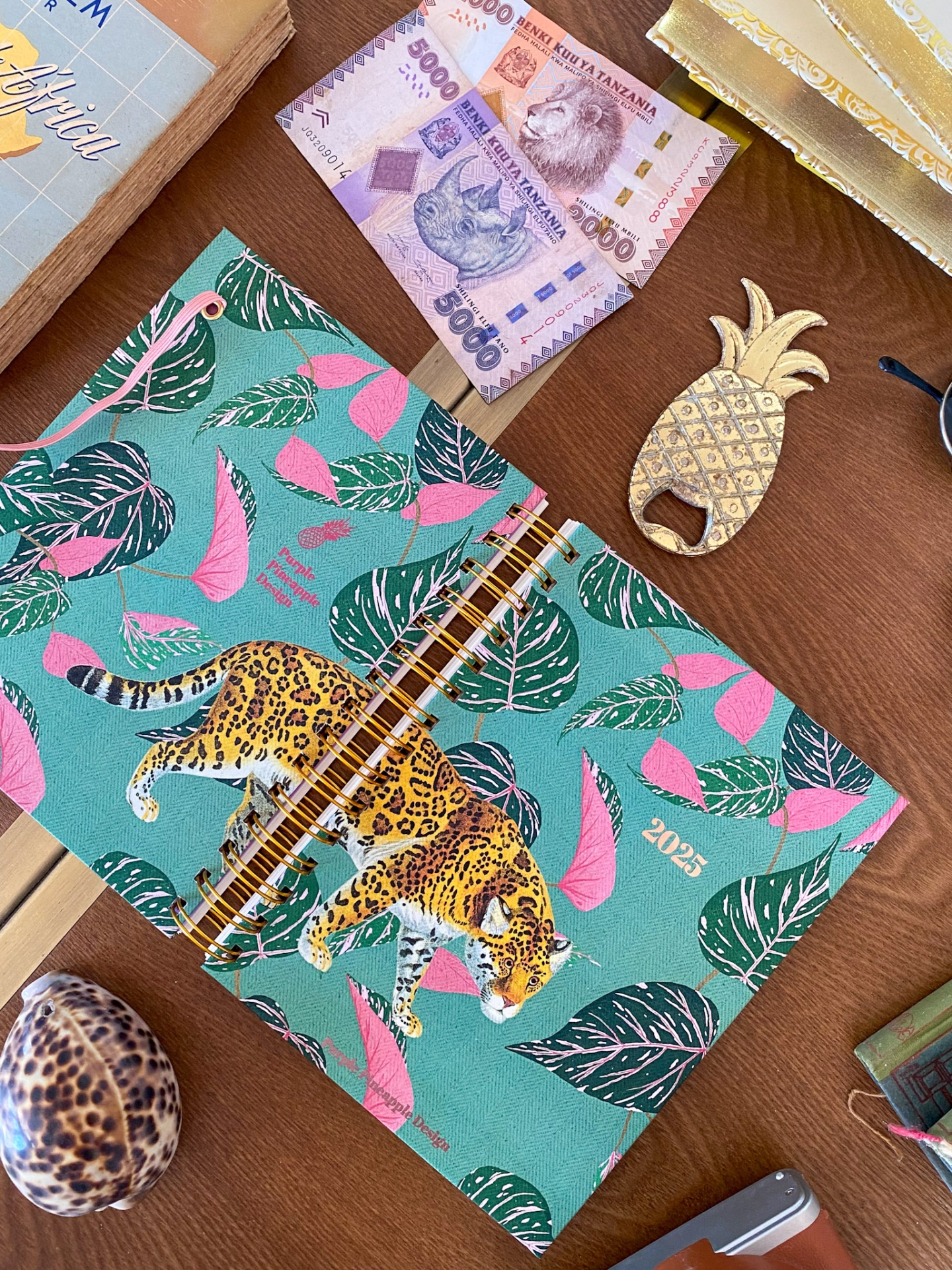 Caderno verde com padrão de folhas e leopardo, dinheiro, abridor de garrafa ananás, moeda e concha numa mesa.