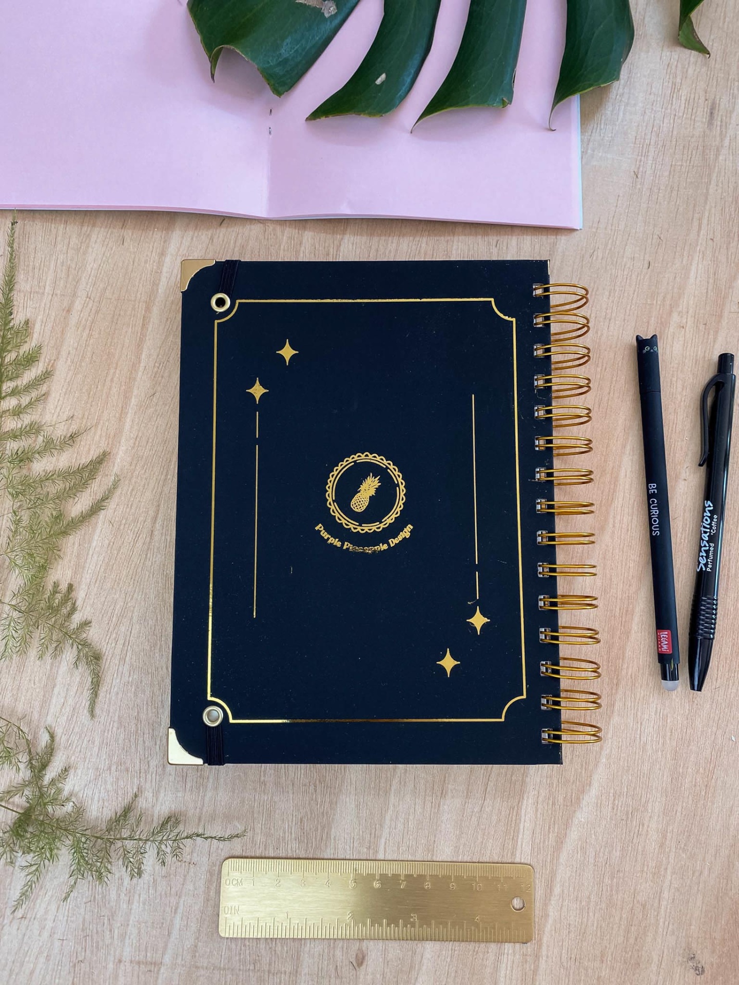 Caderno preto com detalhes dourados e acessórios de escrita sobre mesa de madeira