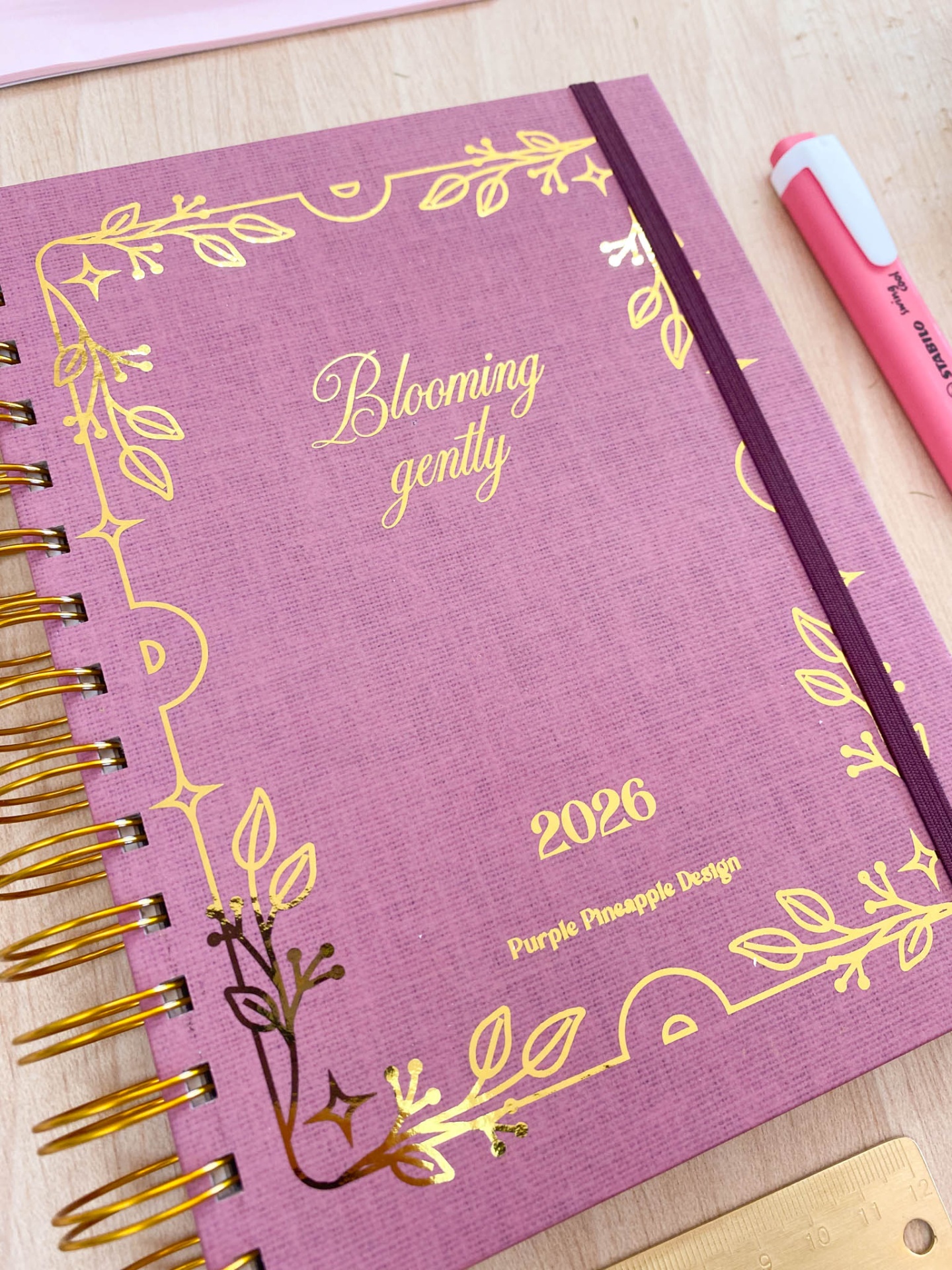 Caderno rosa com detalhes dourados e texto 'Blooming gently' e '2026 Purple Pineapple Design'