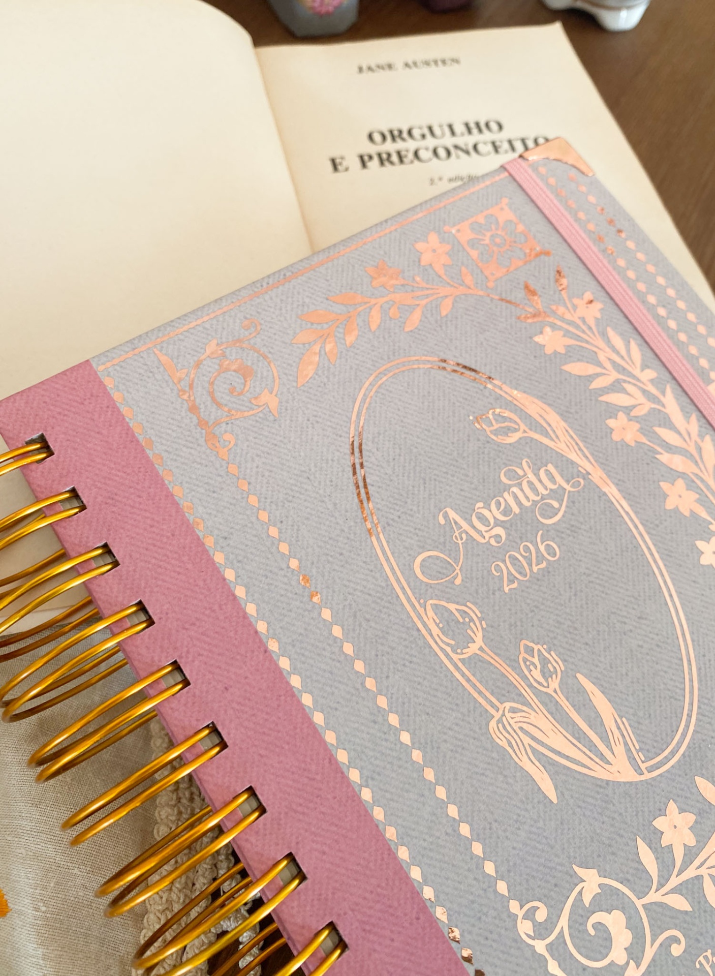 Agenda 2026 com capa cinza e rosa com detalhes dourados floral, sobre livro aberto de Jane Austen