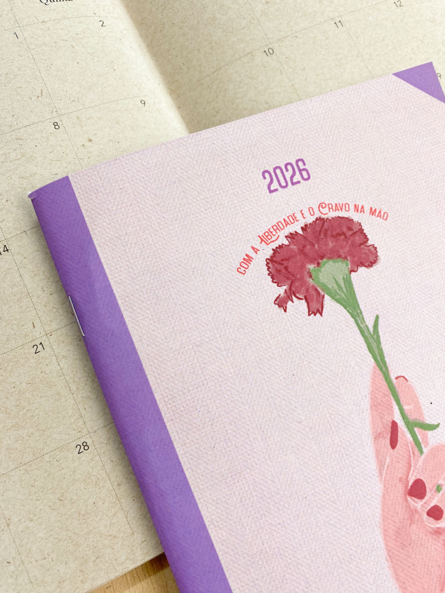 Agenda 2026 com capa lilás e imagem de mão a segurar cravo vermelho sobre calendário