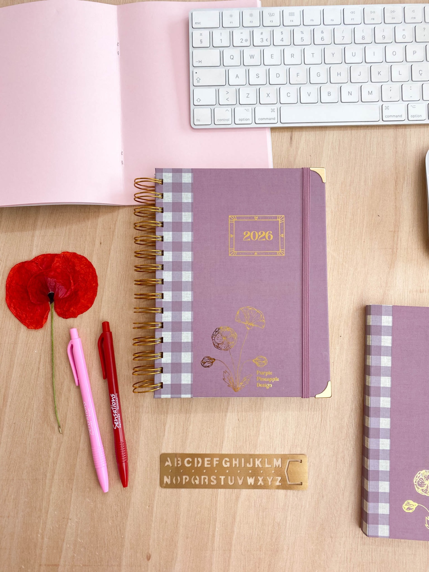 Caderno lilás com padrão xadrez, canetas, flor e teclado sobre mesa de madeira