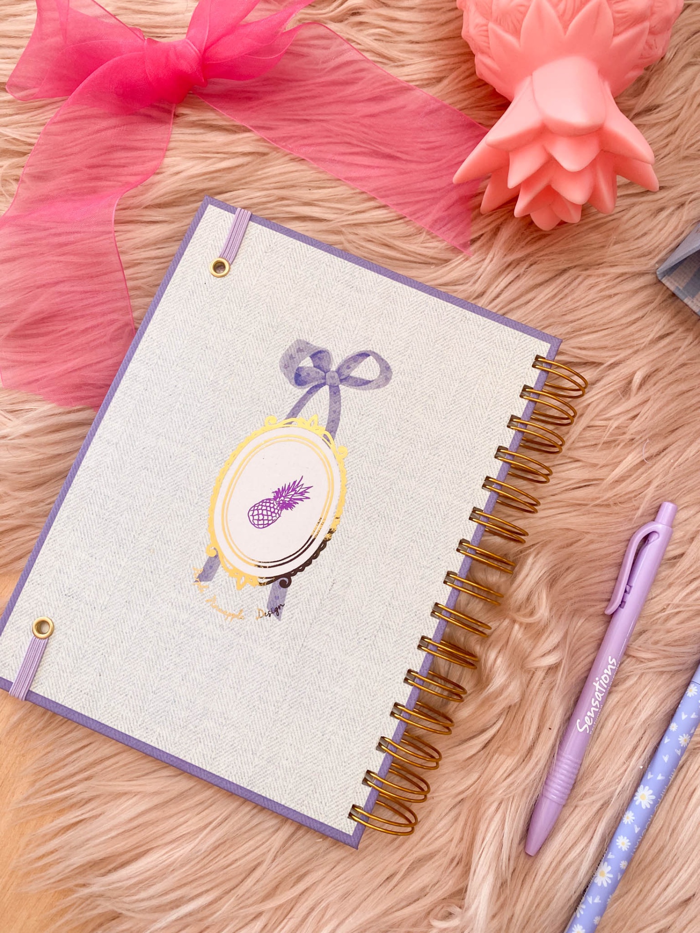 Caderno com capa bege e lilás com ilustração de ananás, caneta lilás e fita rosa sobre superfície felpuda