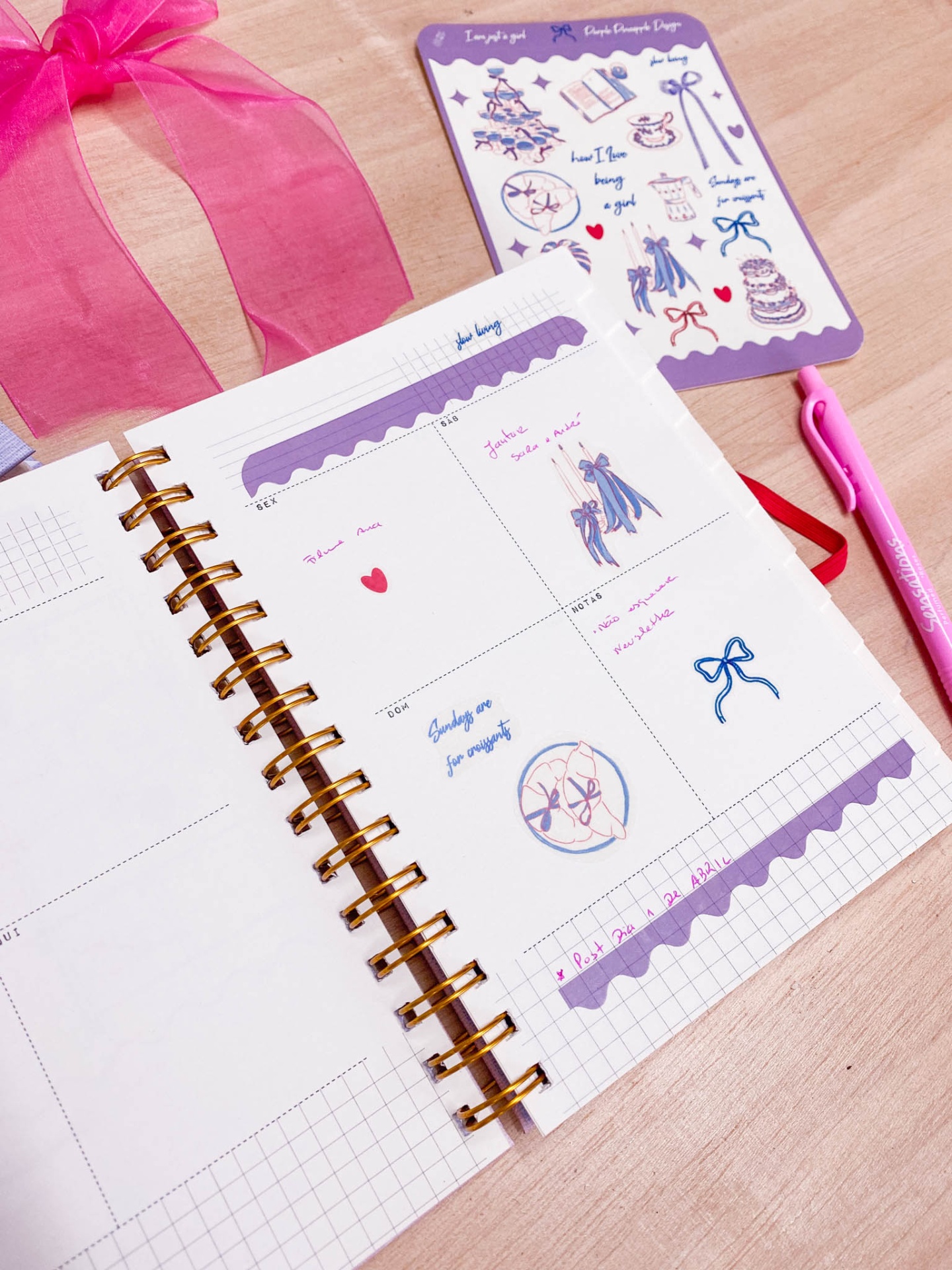 Agenda aberta com adesivos coloridos, caneta rosa e fita de cetim rosa sobre mesa