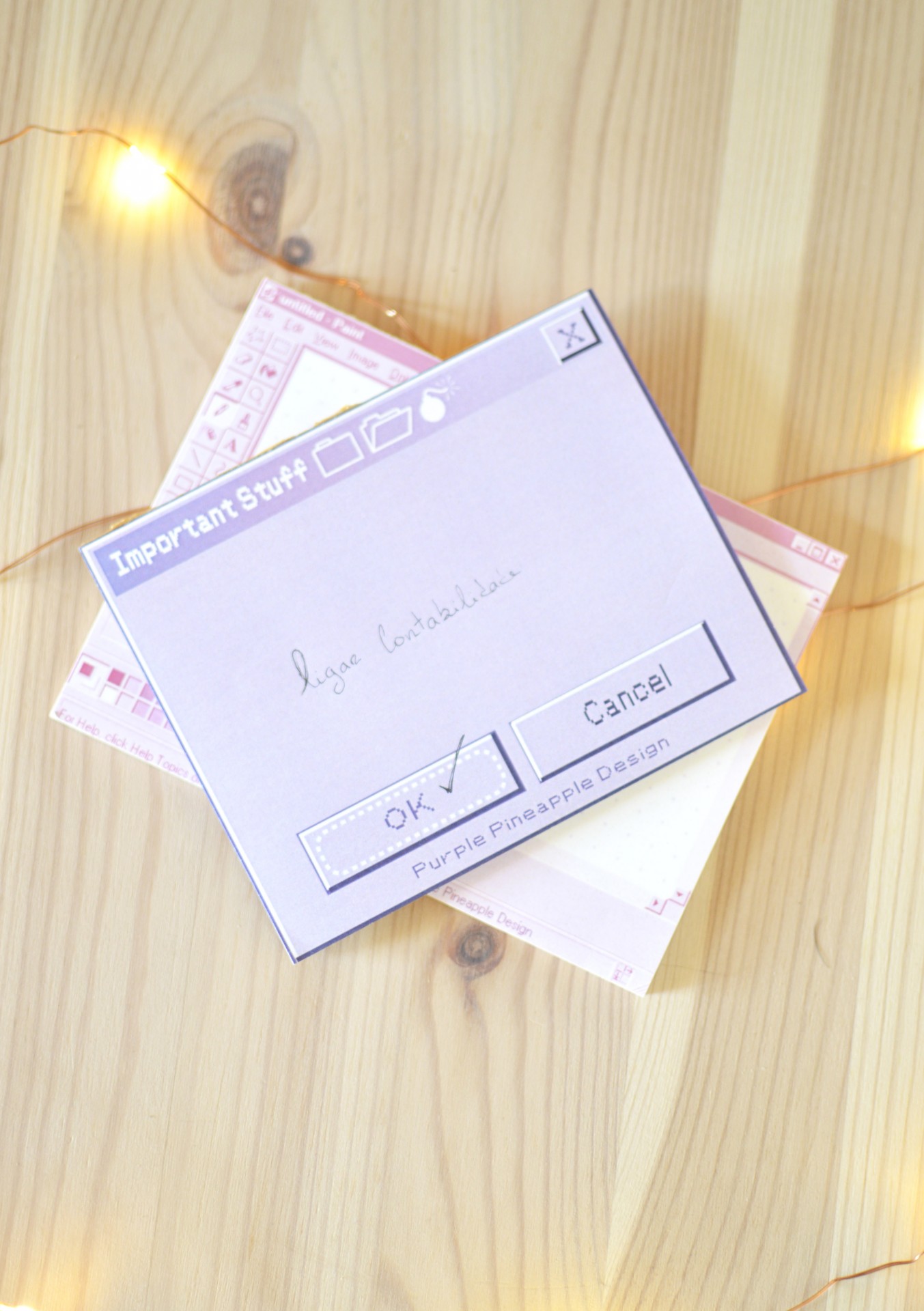 Blocos de notas com design de janelas digitais, lilás e rosa, sobre mesa de madeira clara com luzes de LED.