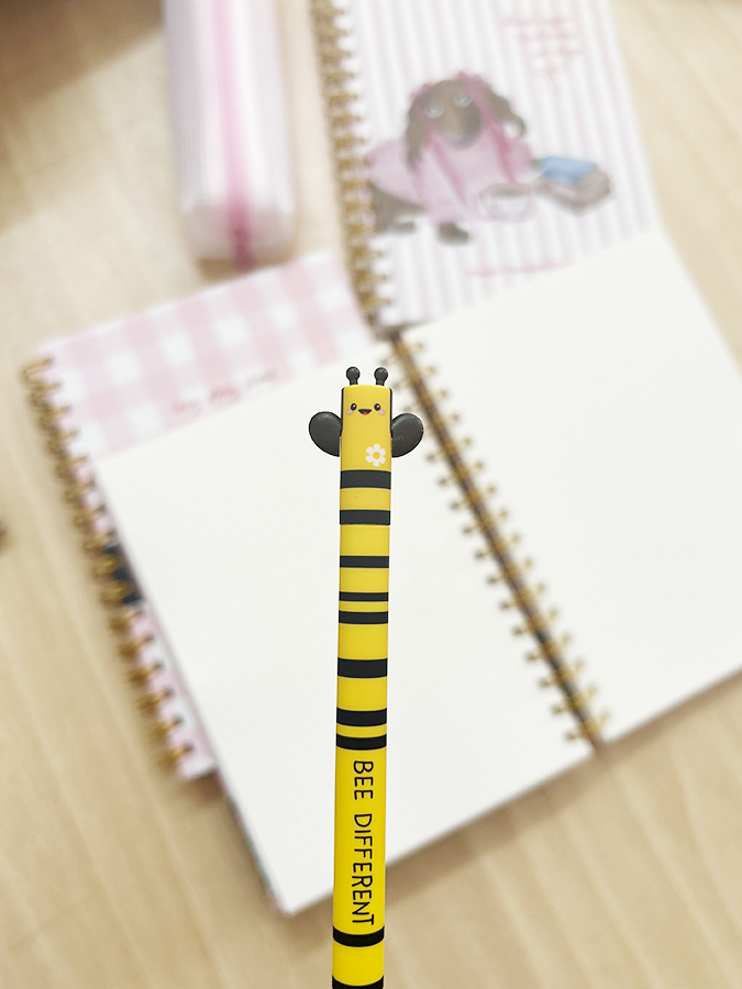 Lápis amarelo e preto com tema de abelha em frente a cadernos espiral com capas rosa e branco