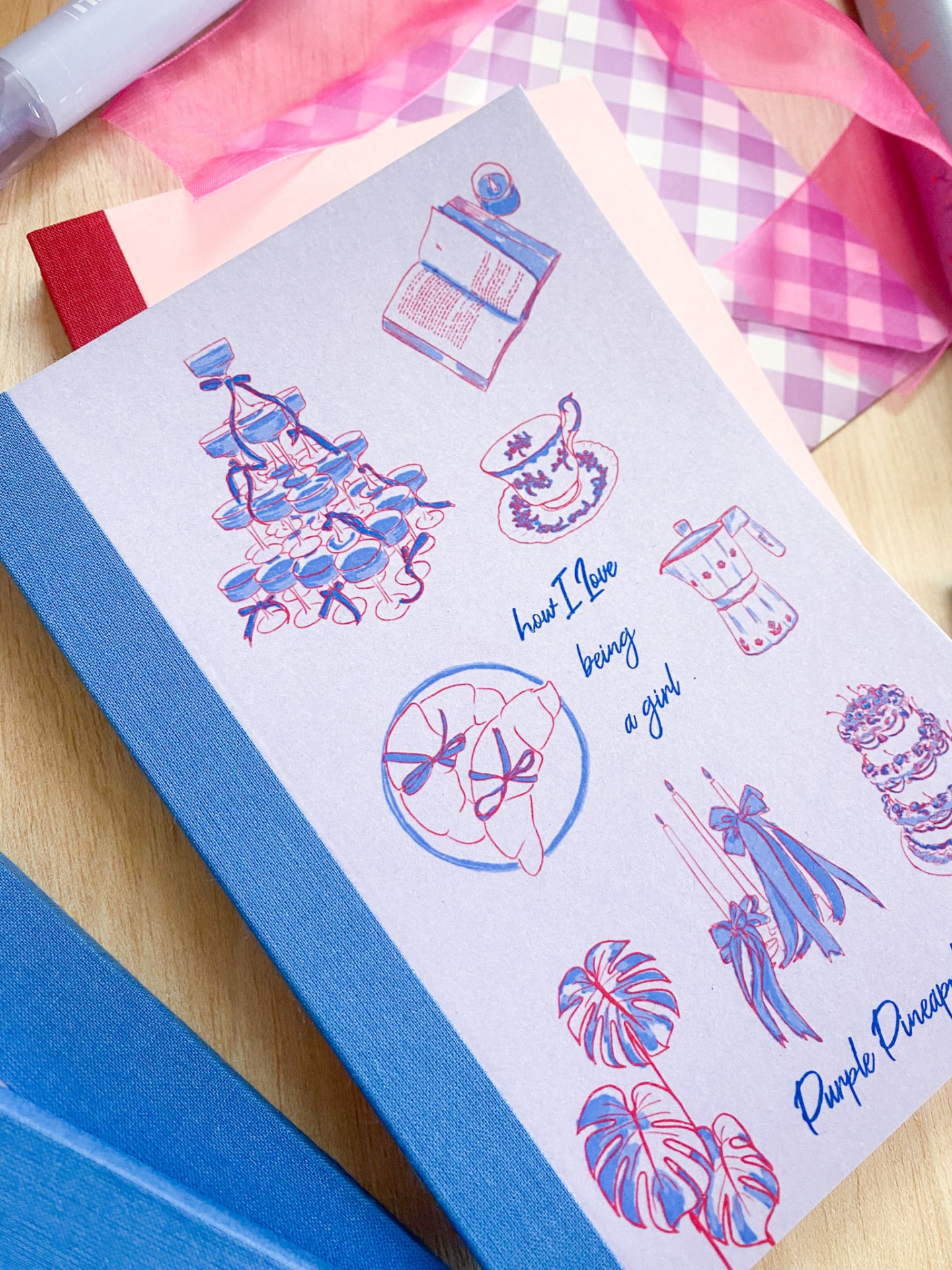 Caderno com capa decorativa com desenhos e textos em azul e rosa, tecido xadrez ao fundo