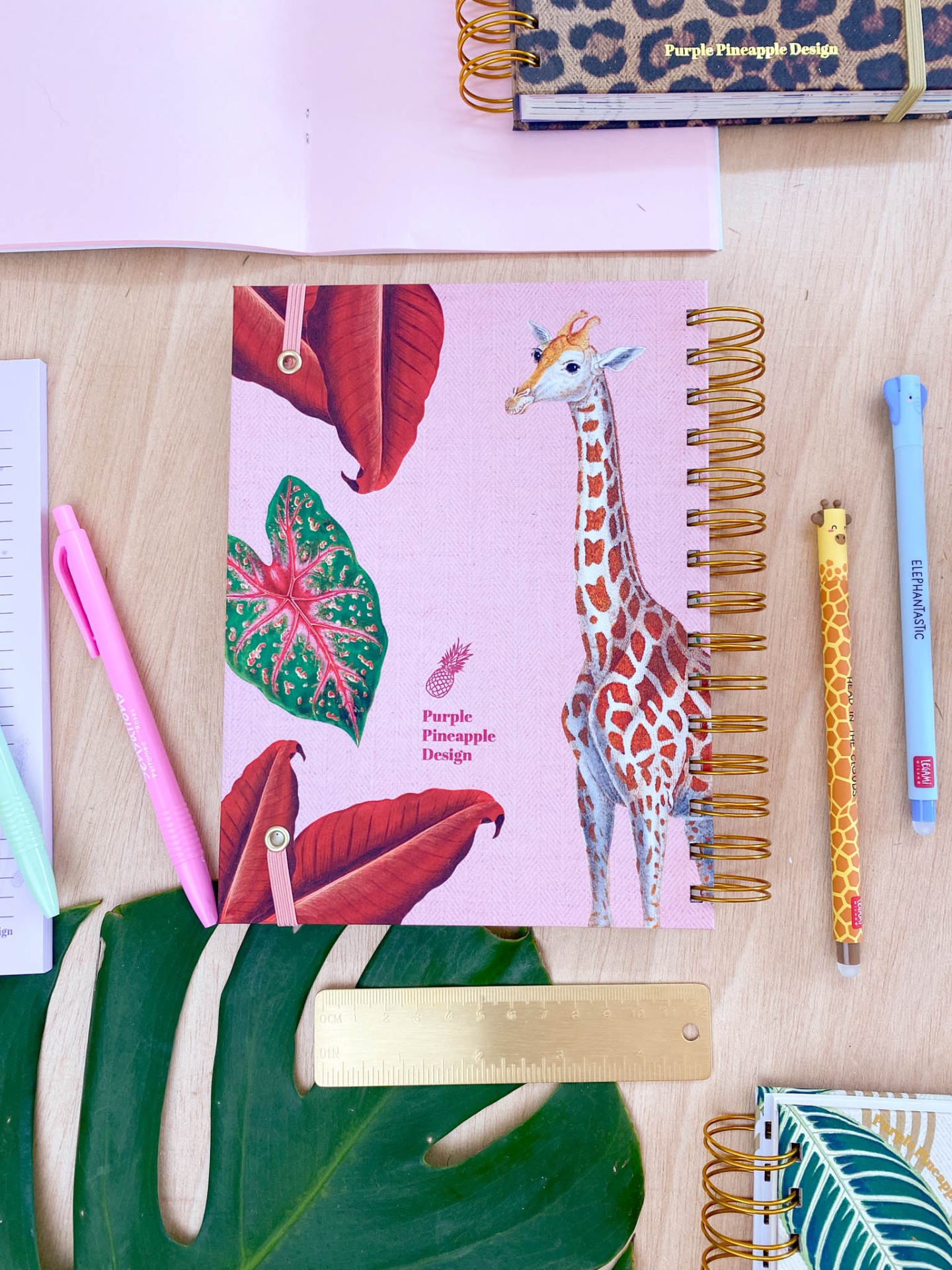 Caderno espiral rosa com ilustração de girafa e folhas, texto Purple Pineapple Design, sobre mesa com materiais escolares