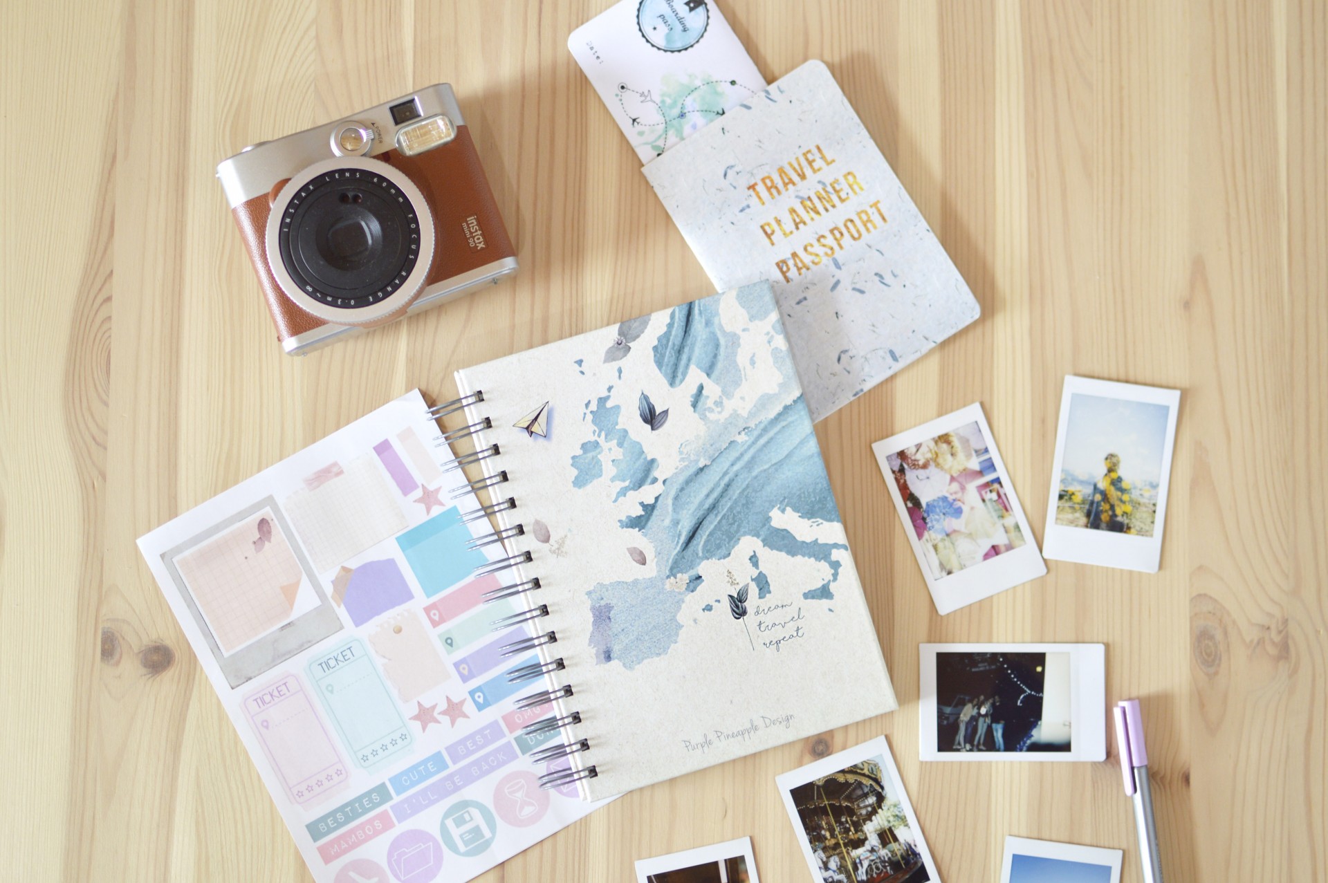 Câmara vintage, caderno de capa azul e bege, autocolantes coloridos, fotografias instantâneas e passaporte sobre madeira clara