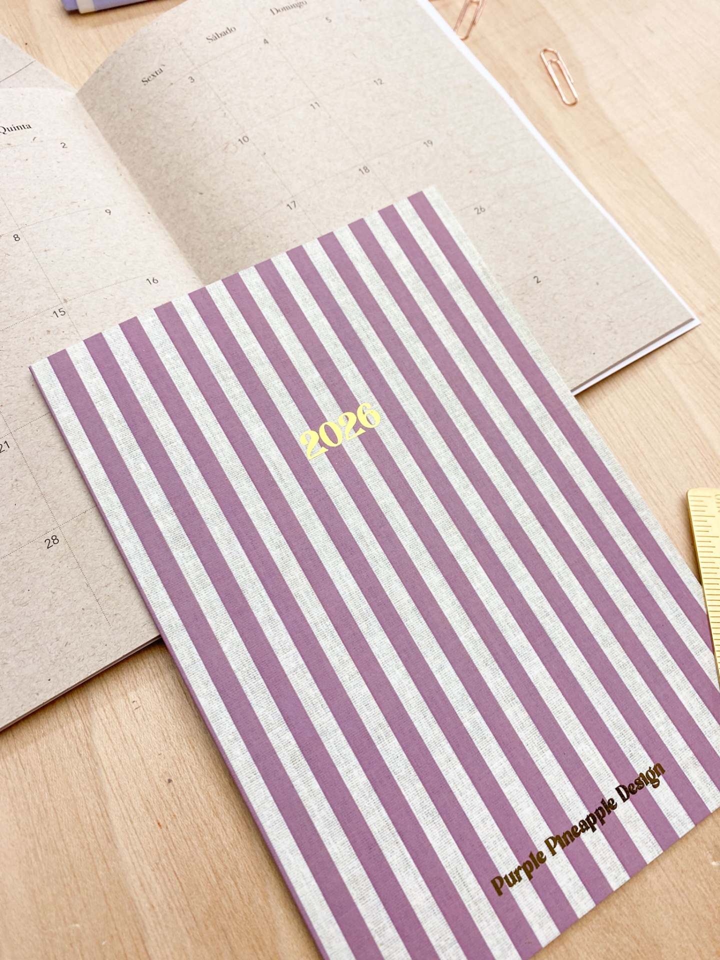 Agenda 2026 riscada lilás e branca com texto dourado sobre mesa de madeira.