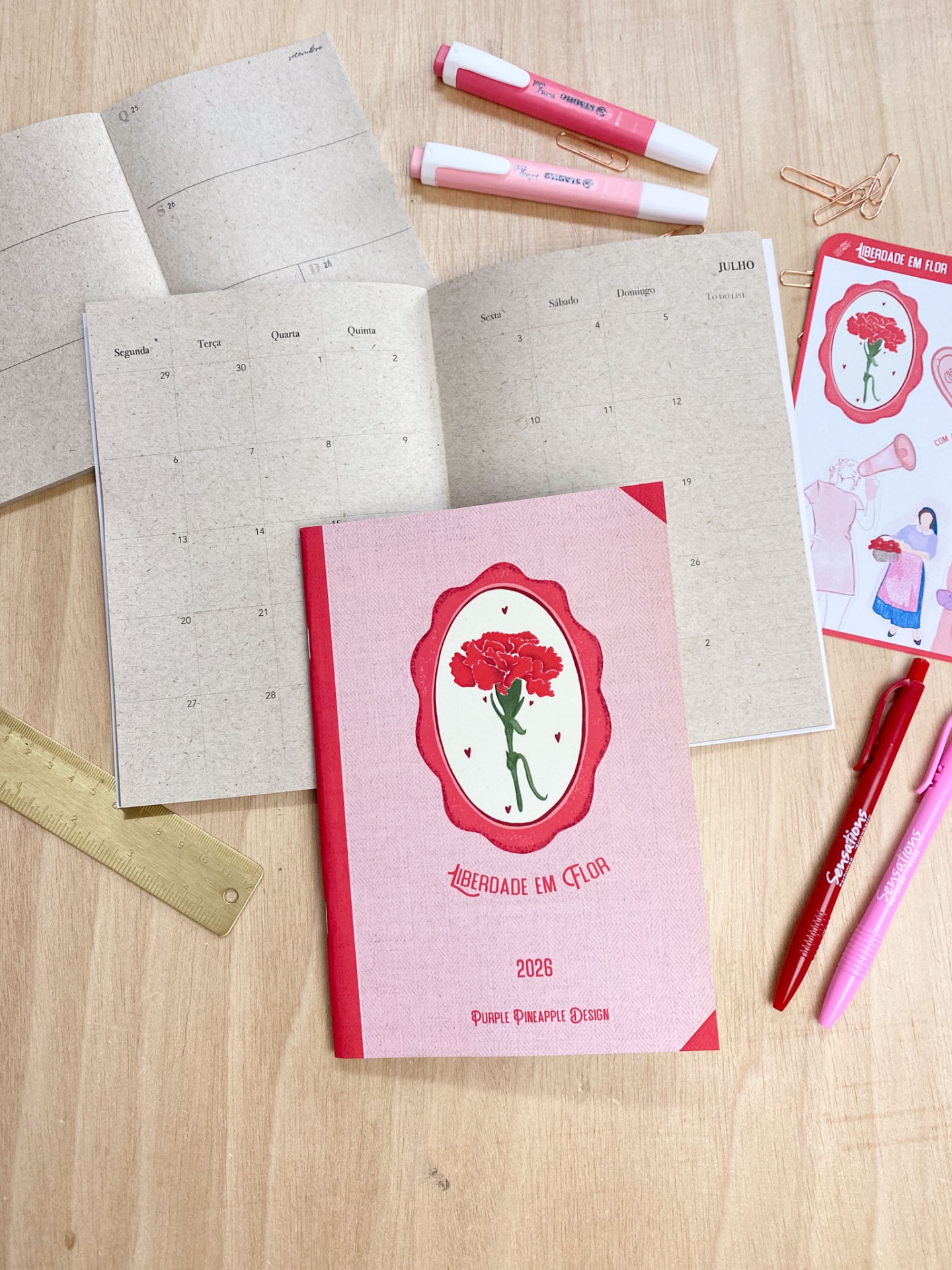 Agenda 2028 rosa com desenho de flor e texto LIBERDADE EM FLOR, aberta, com marcadores e canetas numa mesa de madeira.