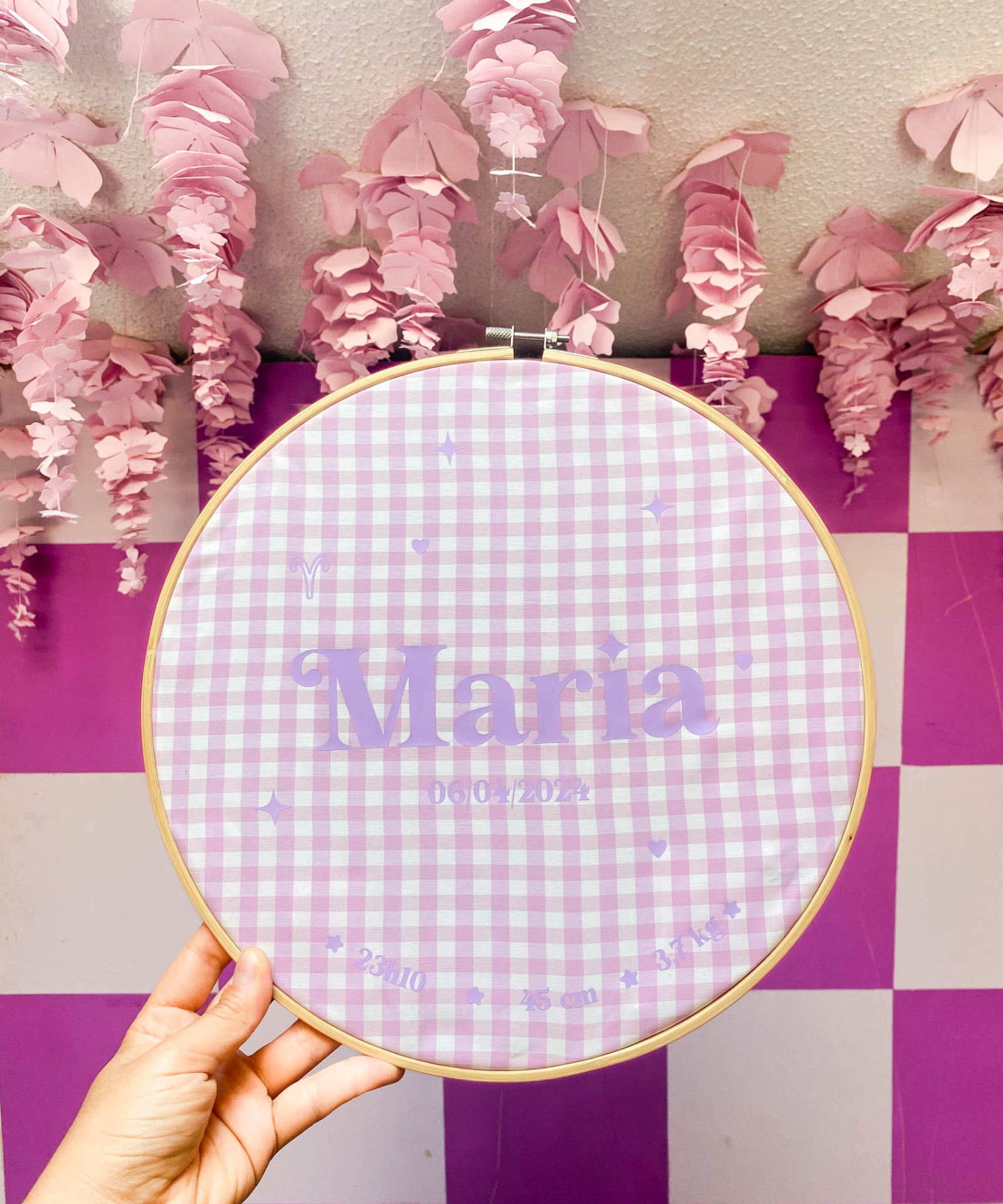 Bastidor com tecido xadrez lilás e branco com nome e data, fundo quadriculado roxo e branco com flores cor de rosa