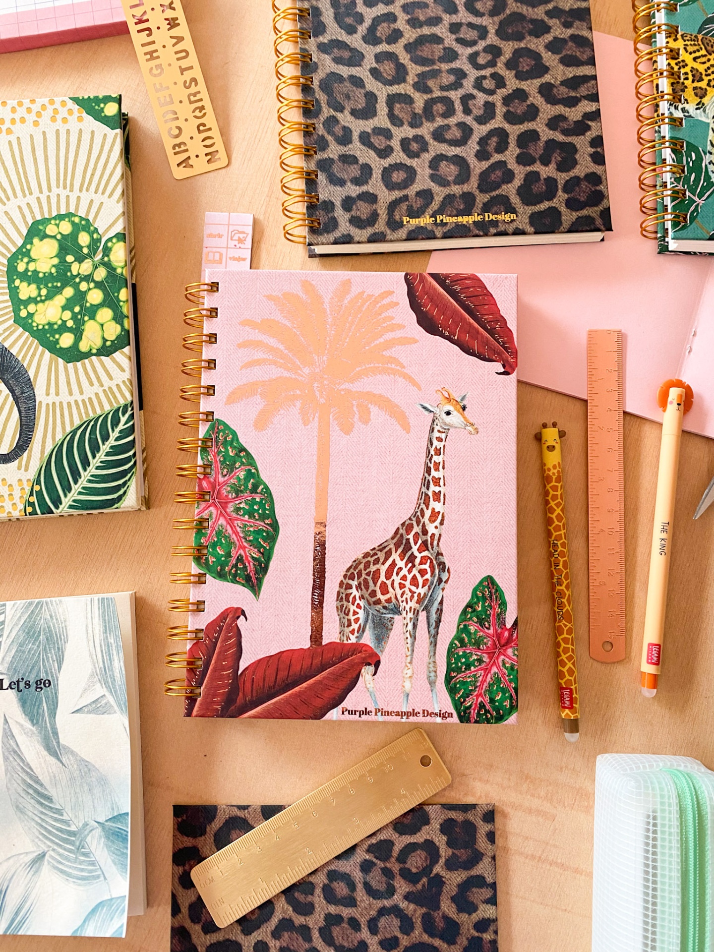 Cadernos decorativos espirais com padrões animais e tropicais ao lado de réguas e canetas numa mesa.